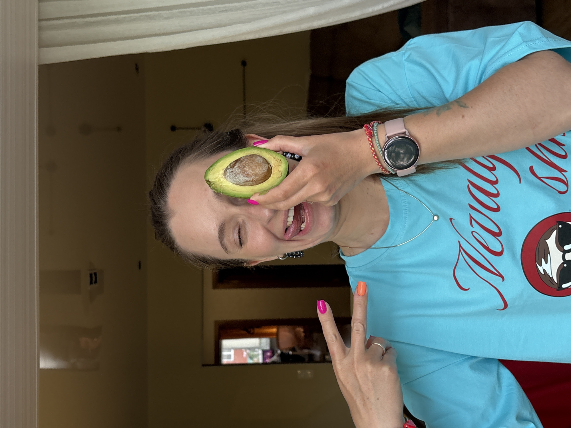 A woman is pictured inside a building, with a window providing a view of another building. She is holding half of an avocado over her left eye, smiling, and giving the peace sign with her right hand. She is wearing a light blue t-shirt with a red design, a watch, and beaded bracelets on her left wrist. Her nails are painted in different colors. She is wearing jewelry and earrings. The location cannot be determined to be Birobidzhan, Russia based on the provided image.