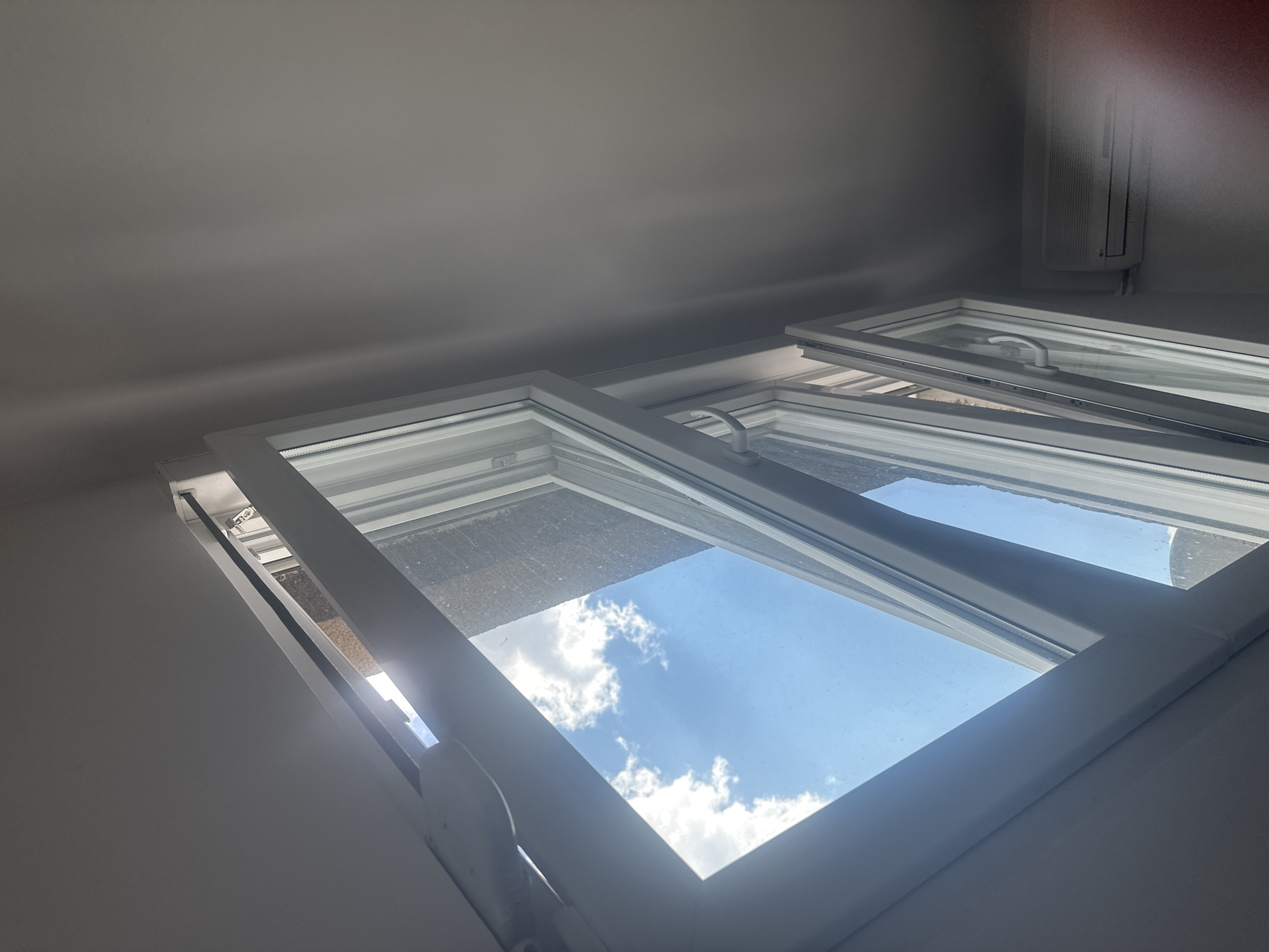 This media file captures an upward-angle view of an open white window frame, revealing a bright blue sky with scattered white clouds. Sunlight streams in from the left, creating a lens flare and illuminating parts of the window and the surrounding white wall. The window appears to be a modern, tilt-and-turn style with a white handle visible on the lower pane. The top pane is tilted inwards, while the lower pane is also slightly ajar. The interior wall is painted a light, neutral color, and a hint of what appears to be an air conditioning unit is visible on the right side of the frame in the background. The overall impression is one of a sunny day, likely during the daytime, given the intensity of the sunlight and the clear blue sky. The setting appears to be indoors, looking out through a window. There is no visible text in the image.