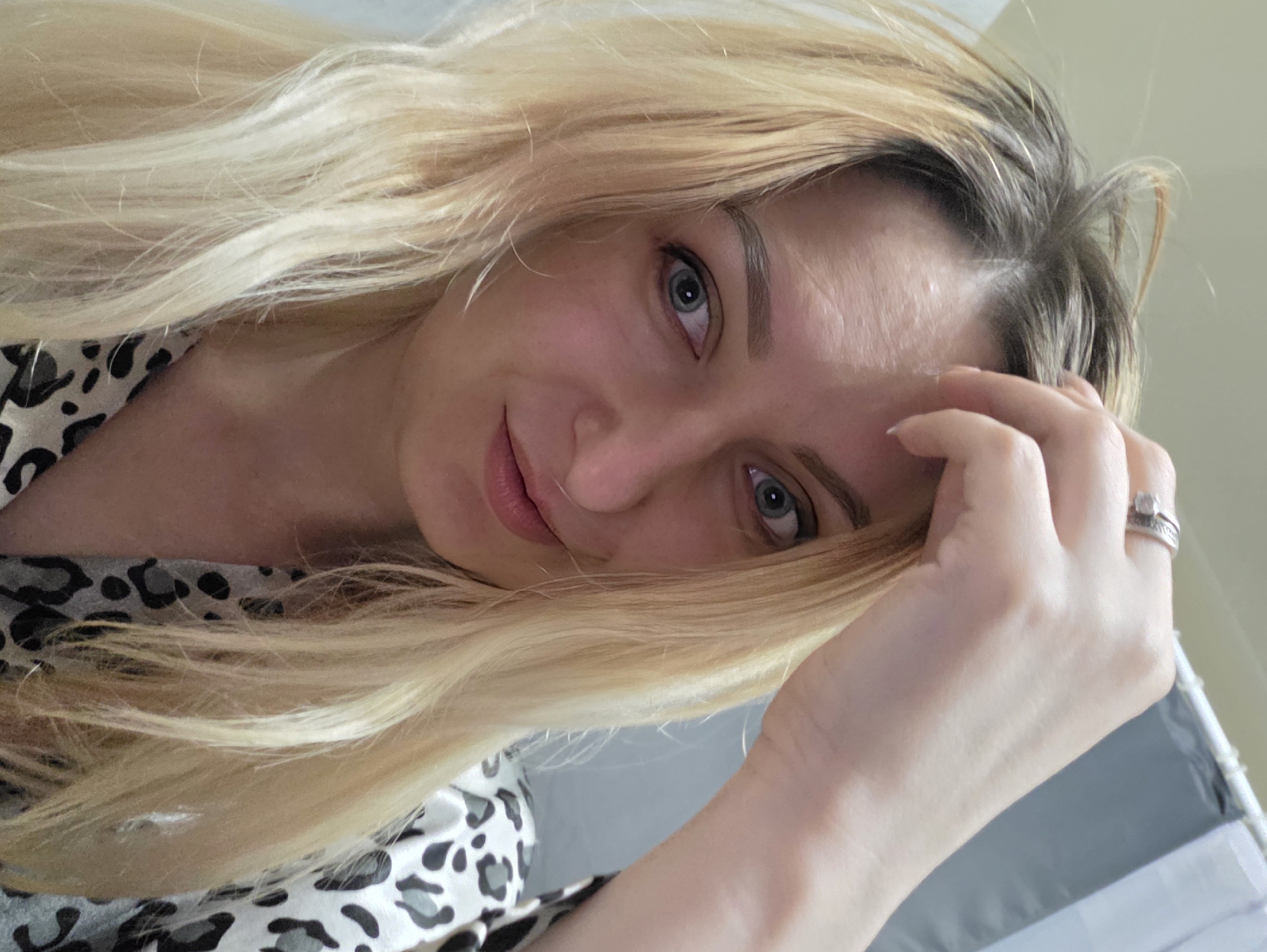 An adult female, appearing to be in her 20s or 30s, is captured in a close-up, head-and-shoulders portrait in the city of Minsk, Belarus. She has long, blonde, wavy hair with discernible darker roots at the scalp. Her light blue eyes are directed at the viewer, accompanied by a subtle, closed-mouth smile. Her right hand is raised, with fingers resting on her forehead near the hairline. A silver-toned ring with visible detailing is present on the ring finger of her right hand. She is wearing a garment featuring a grey, black, and white leopard print pattern. The background consists of a light-colored wall or surface on the left and a grey fabric, possibly a curtain on a rod, on the upper right side of the frame, suggesting an indoor, domestic setting.