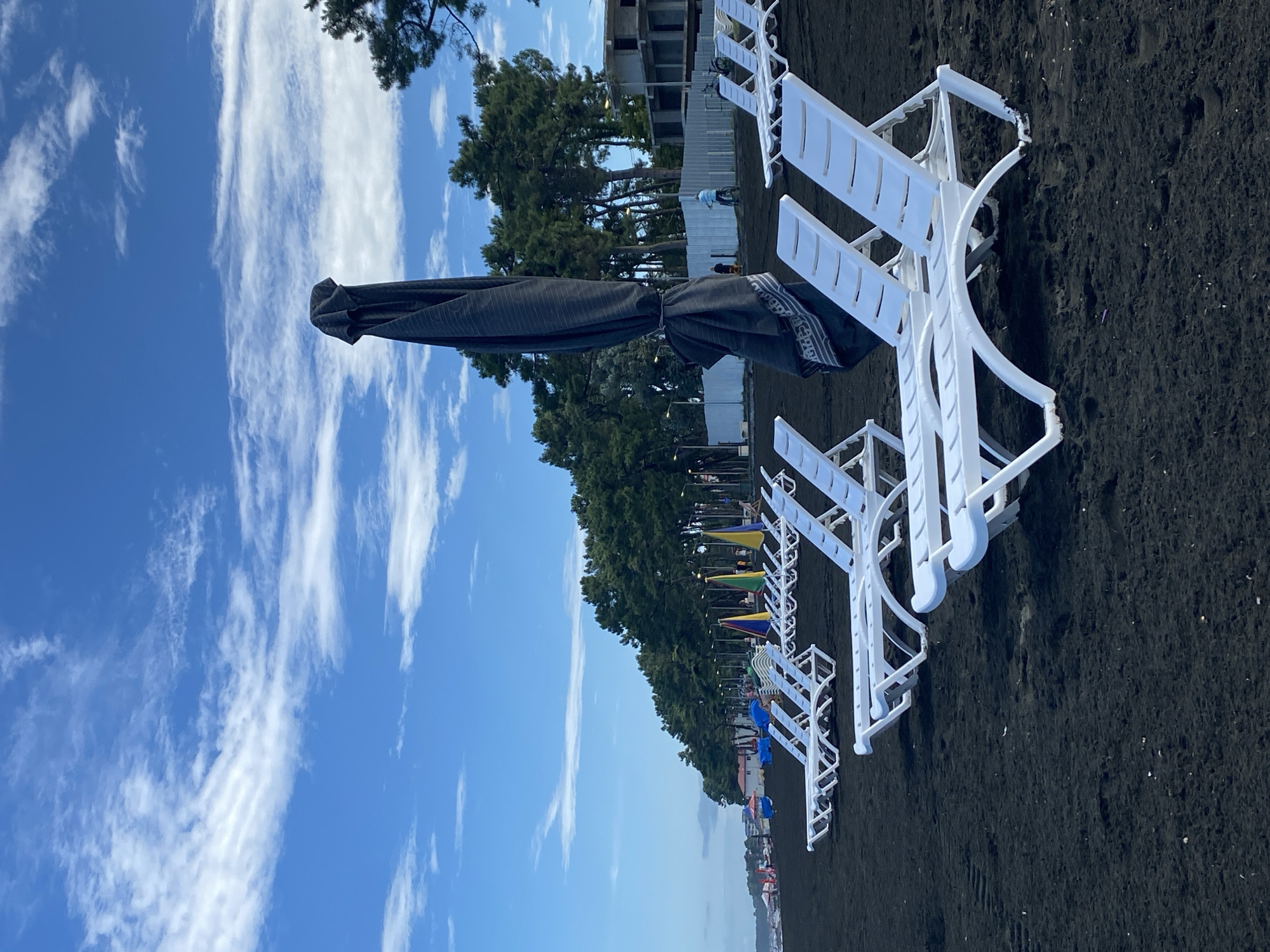 The image depicts a beach scene in Ureki, Georgia, characterized by its distinctive black sand. In the foreground, a row of empty white plastic lounge chairs is neatly arranged on the dark sand. One large, dark-colored beach umbrella is closed and tied, standing upright in the center of the frame. Further back, more lounge chairs and colorful umbrellas are visible, hinting at a recreational area. A line of lush green trees forms a backdrop to the beach, with a glimpse of a building under construction and a white fence in the distance. The sky above is a clear blue, punctuated by wispy white clouds. A person on a bicycle is visible in the middle ground, near the fence, and a few other individuals can be seen in the background near some umbrellas.