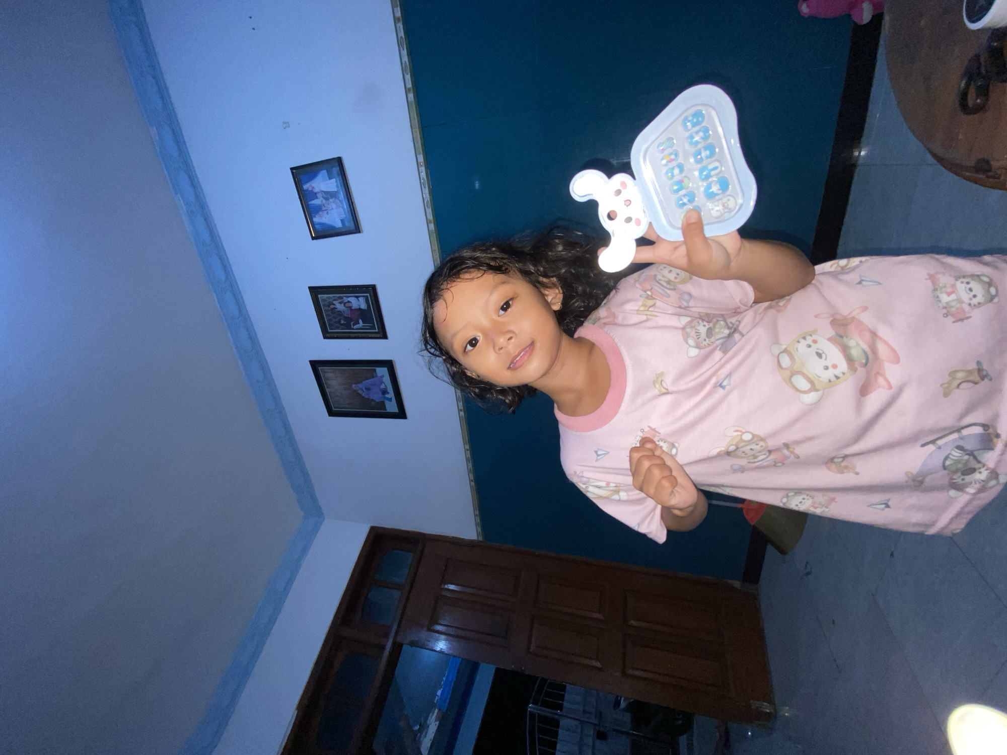 A young girl with dark, curly hair is holding a light blue and white toy phone in her right hand. She is wearing a pink, patterned pajama top. Three framed pictures are visible on the wall behind her. A dark wooden door is located in the lower part of the frame. The location is Gelam, Indonesia.