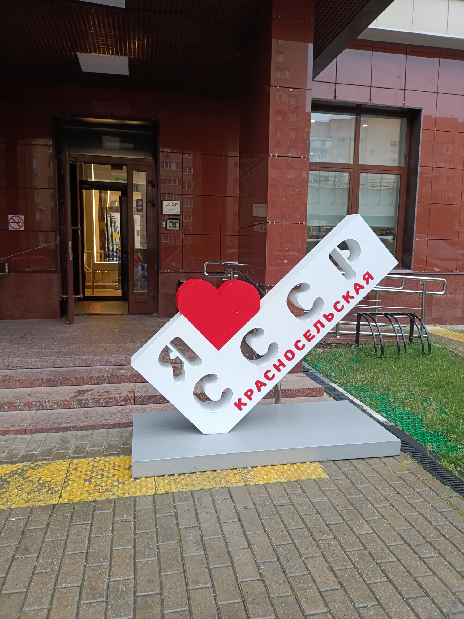 A sign with a red heart and white letters is placed on a paved area in front of a building. The sign reads "Я люблю СССР" (I love USSR) and also has the word "КРАСНОСЕЛЬСКАЯ" (Krasnoselskaya) written in red on a white slanted plank. The building is made of red brick and has large windows. An open doorway reveals a well-lit interior. The paved area is bordered by a yellow tactile paving strip and a grassy area with a black bicycle rack. The scene appears to be in an urban environment.