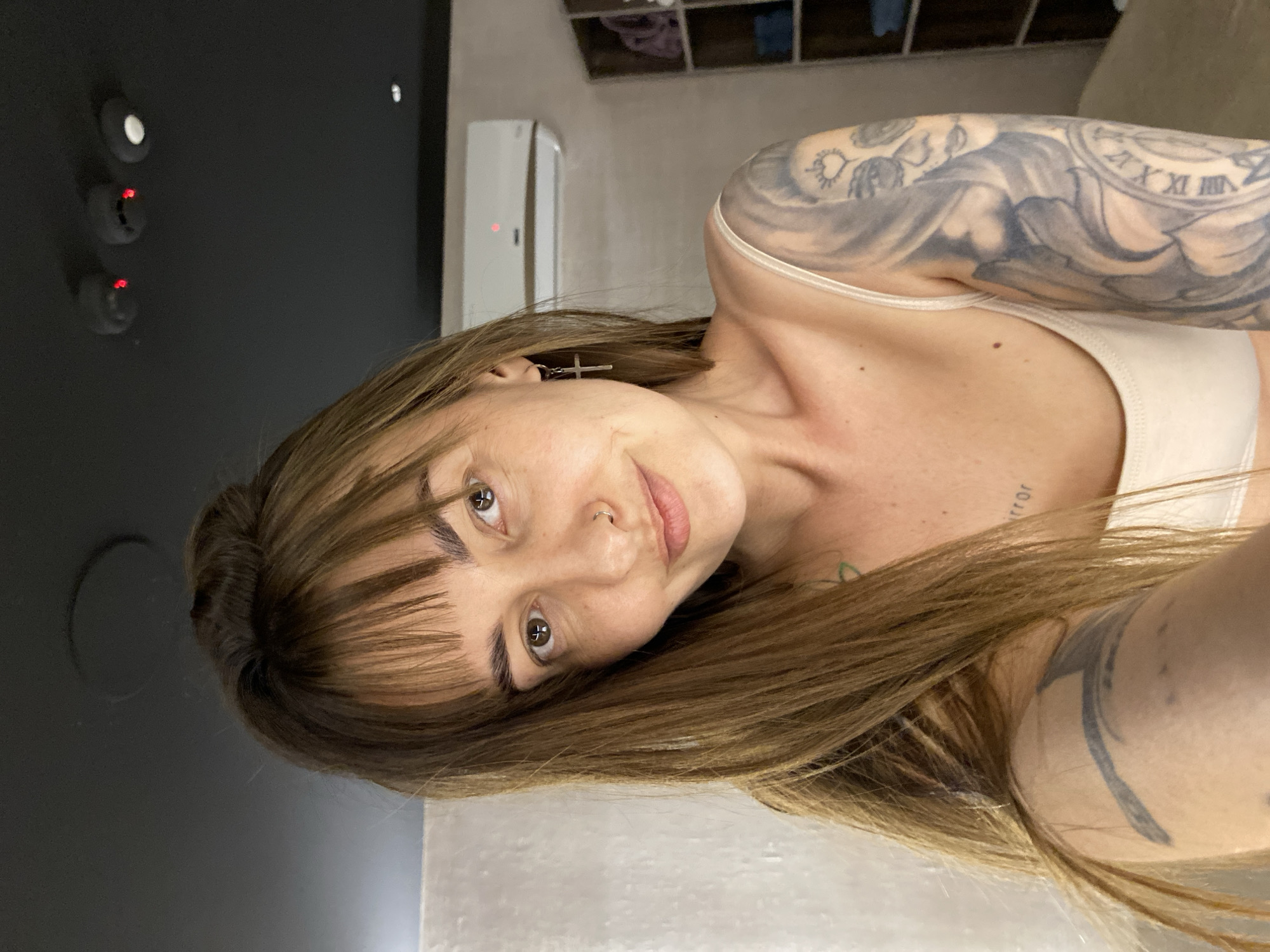 A woman with brown hair and bangs is posing for a selfie. She has a nose ring and a cross earring. Tattoos are visible on her left arm and chest. She is wearing a beige top. The background includes a dark gray wall with control panel and a beige wall with a white air conditioning unit. An open closet with clothes inside is visible at the top of the frame. No specific location in Votkinsk, Russia, is identifiable from the image.