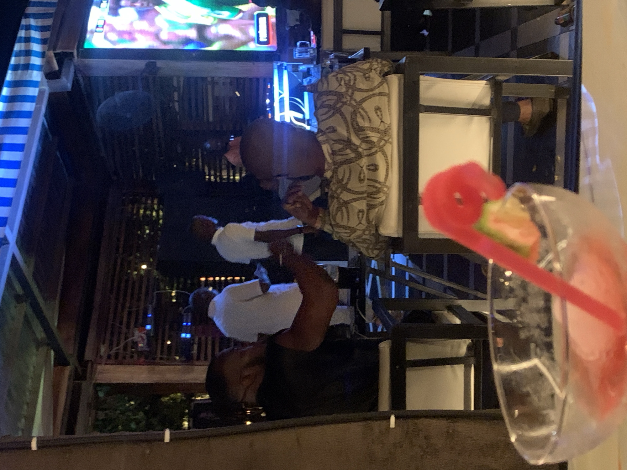The image depicts an indoor-outdoor seating area, possibly a bar or restaurant, in Abuja, Nigeria. In the foreground, a glass of pink beverage with a red straw and a slice of fruit is partially visible. The scene captures several individuals, some seated and others standing, engaged in various activities.
To the left, a man with a black sleeveless shirt is seated, facing away from the camera, with his left arm resting on the back of his chair. A glass is placed on a table in front of him. In the mid-ground, three men are visible. Two of them, wearing white shirts, are standing and appear to be in conversation. Another man, seated and facing the screen, is wearing a patterned shirt.
In the background, a large television screen displays colorful images, and a blue light emanates from a bar area. The environment suggests a lively social setting, with ambient lighting and furniture indicating an establishment designed for patrons to relax and socialize. The overall impression is of an evening gathering at a venue in Abuja.