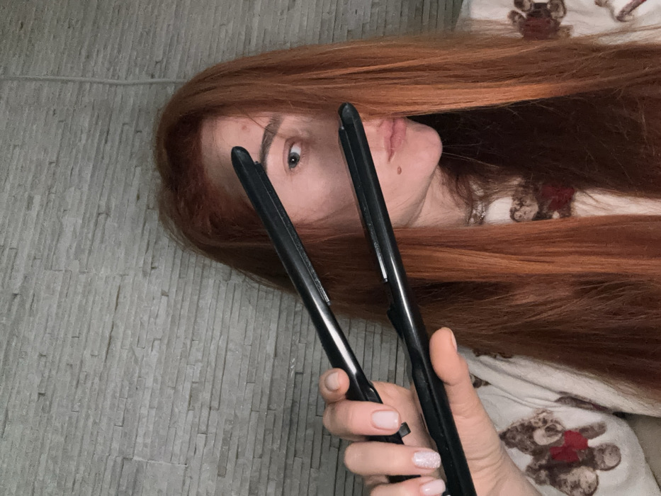 A close-up image taken in Budva, Montenegro, depicts a female individual holding a black hair straightener. The individual's long, reddish-brown hair is partially draped over the right side of her face, obscuring part of her nose and eye. Her left eye, light blue in color, is visible. A small blemish is present on her forehead, and a mole is visible below the left corner of her mouth. Her left hand, with manicured nails painted in a light pink/nude shade (the ring finger featuring glitter), grips the hair straightener, holding its plates open. She is wearing a light-colored garment patterned with teddy bears, one of which displays a red heart. The background consists of a light grey wall with a textured, stacked stone or brick pattern.