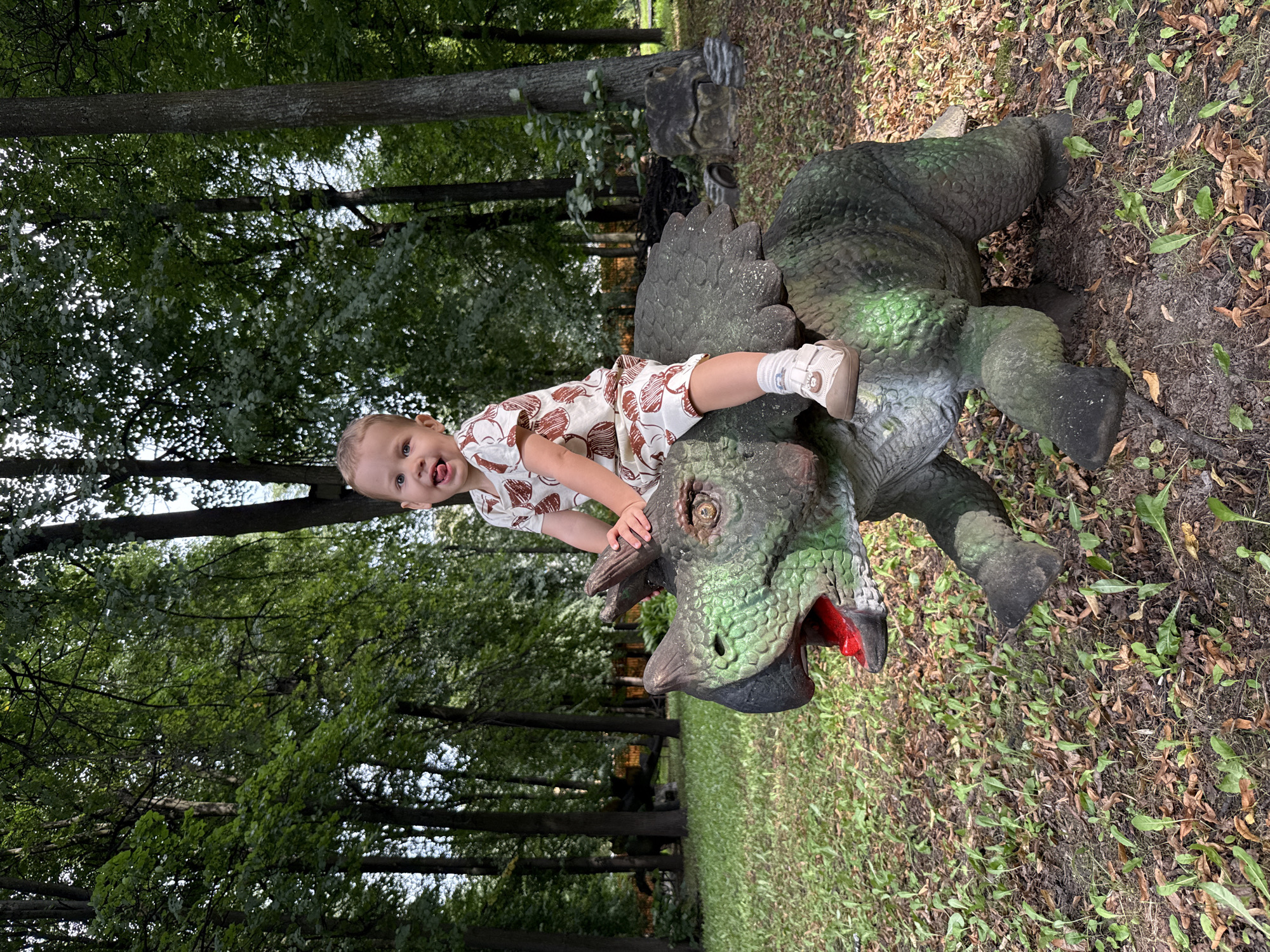In the city of Rusanovo, Russia, a young child, possibly a toddler, is pictured sitting atop a green and black dinosaur sculpture. The child is wearing a light-colored onesie with a repeating brown circular pattern and white socks with light brown shoes. They are looking towards the camera with their tongue sticking out. The dinosaur sculpture, which appears to be made of concrete or a similar material, is positioned in a grassy area with fallen leaves. Behind the sculpture, a dense forest with tall trees forms the background. The lighting suggests it is daytime, with sunlight filtering through the foliage. The overall scene depicts a playful moment in an outdoor, possibly park-like, environment.