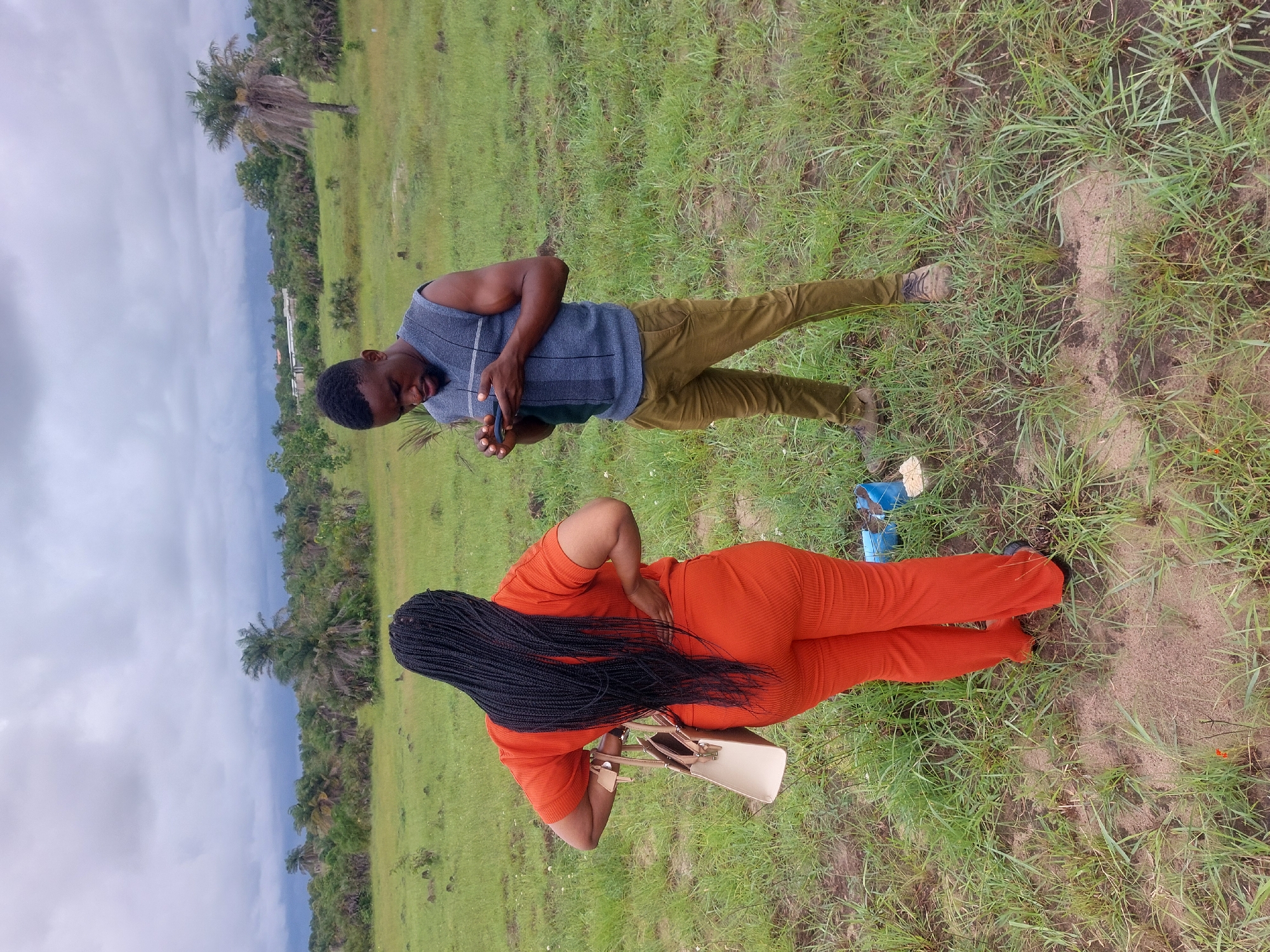 Two people are standing in a grassy field under an overcast sky. A man is facing downward, looking at a small object in his hands. He is wearing a grey sleeveless shirt and brown pants. A woman in orange is standing with her back to the camera, her long black braids falling down her back. She is carrying a light-colored purse. The location is Dar es-Salaam, Tanzania. Tall trees are visible in the background.