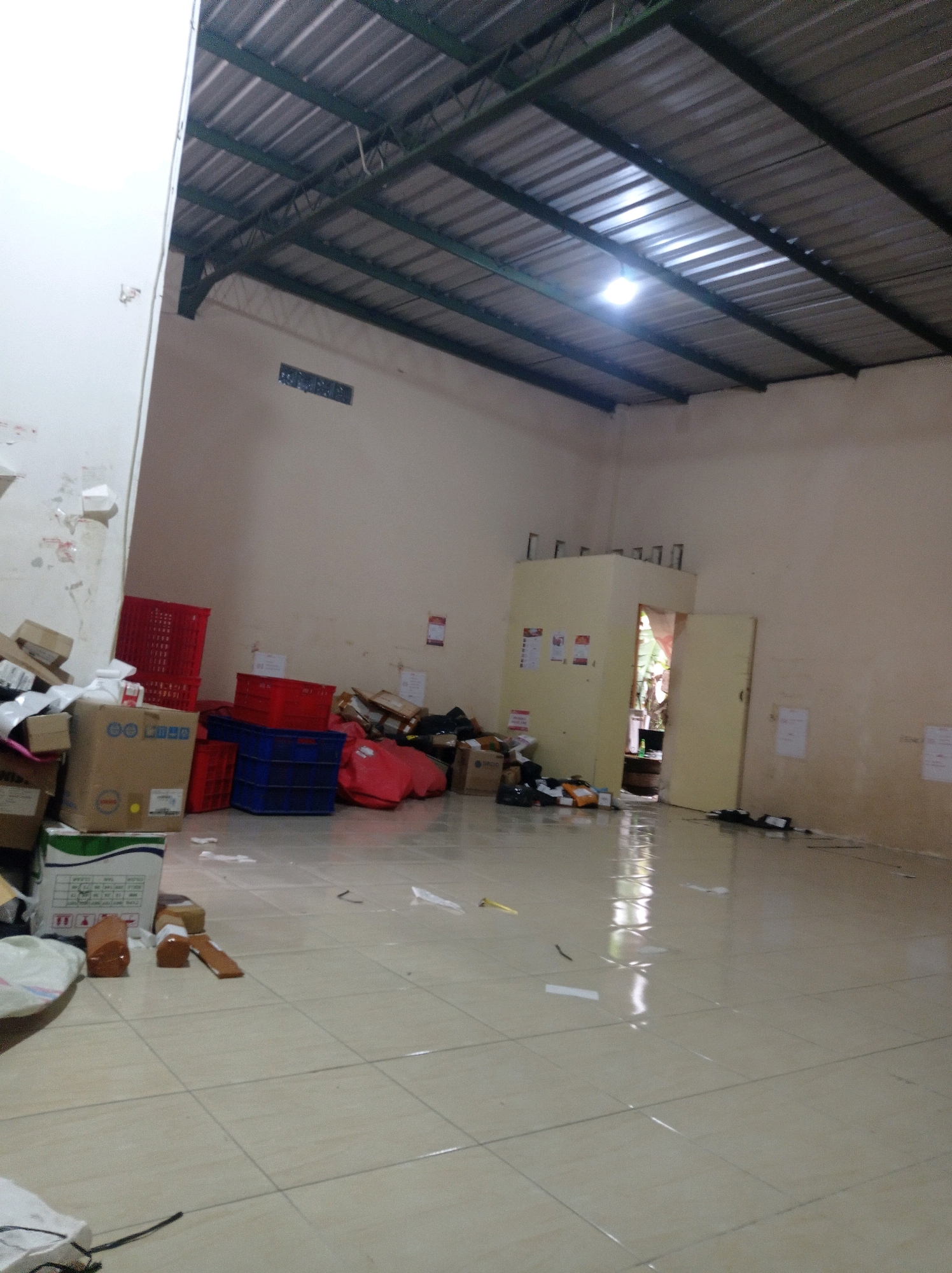The image depicts an interior view of a warehouse or storage room located in Jetis, Indonesia. The room has a high, corrugated metal roof supported by a green metal frame. A single light bulb illuminates the space, casting reflections on the tiled floor, which appears wet or recently cleaned. The walls are painted a light, neutral color and show some minor staining or marks.
The scene is filled with various objects, predominantly boxes and crates, suggesting a distribution or logistics facility. In the foreground and mid-ground, stacks of cardboard boxes of different sizes are visible. Some are sealed, while others are open. There are also several red and blue plastic crates, some filled and others empty, arranged in piles. To the right, a large red bag is present, along with more boxes and what appear to be packages wrapped in brown paper.
Further into the room, there is a partially open doorway leading to another area, which appears to be outdoors or a different interior space with vegetation visible. Adjacent to this doorway is a small, enclosed structure with a light-colored exterior and some posters or notices attached to its walls. Cables and electrical cords are scattered on the floor in the mid-ground and background. The overall impression is one of an active, though possibly cluttered, storage and handling environment. There are no people visible in the image.
