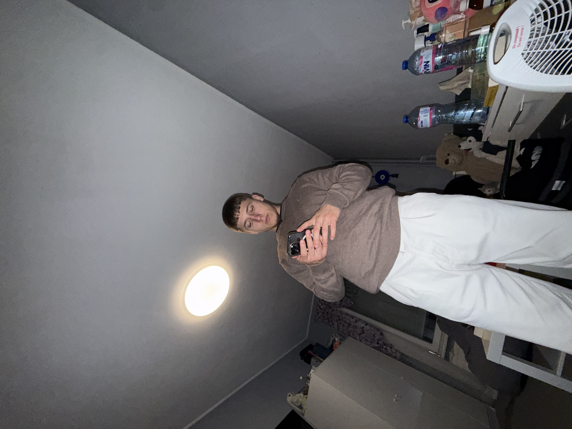 The image captures a young man standing indoors in a casual setting, likely a room in Sofia, Bulgaria. He is seen holding a smartphone in both hands, looking down at the screen, possibly taking a selfie or interacting with the device. He is dressed in a brown long-sleeved sweater and white trousers.
A bright, circular light fixture on the ceiling illuminates the upper part of the room. To the man's right, a cluttered shelf or table is visible, holding various items. Among these are several plastic bottles of "EVIAN" water and a white oscillating fan. Text on the fan is partially visible, reading "НЕ НАКРЫВАТЬ! Do not cover!" Below these items, a brown teddy bear and other stuffed animals are discernible on the shelf. The room's walls are a light, neutral color, and the overall lighting suggests an indoor scene, likely during the evening or night. The man's facial expression is calm and neutral.