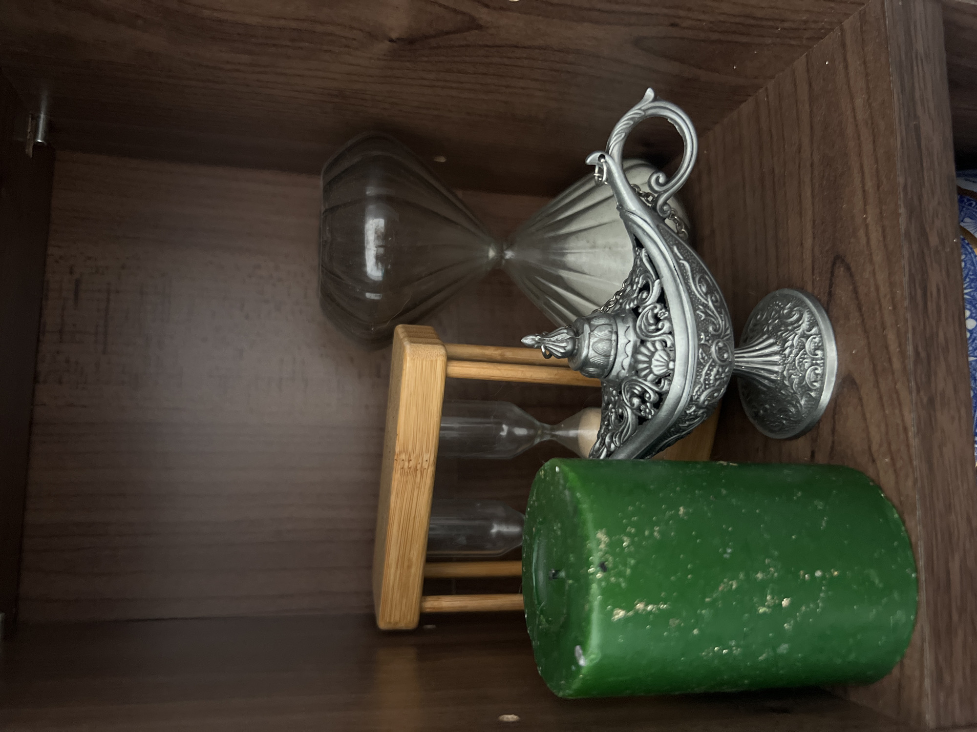 A high-angle view shows the interior of a wooden cabinet or shelf. The dominant objects are an ornate silver-colored metal vessel, two glass hourglasses, a wooden frame, and a green candle. The items appear to be placed randomly, not arranged. The background is the dark brown wood of the cabinet interior. The details required to determine the location in Gorleston-on-Sea, United Kingdom, are not present in the image.