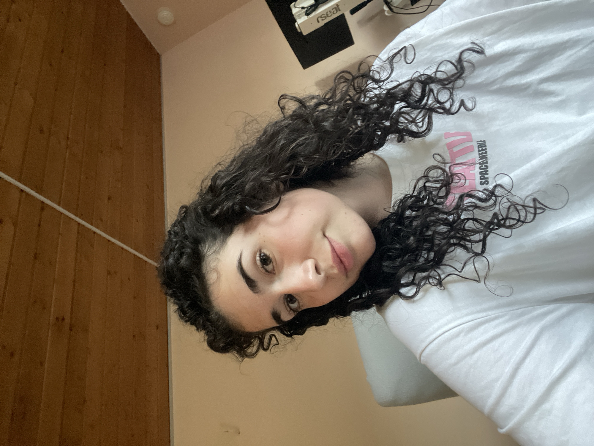 This indoor portrait features a young woman with dark, curly hair looking directly at the camera. Her head is tilted, giving the impression of a selfie or a close-up shot from an elevated angle. She has fair skin, pronounced eyebrows, and appears to be wearing light makeup, with a natural blush on her cheeks and pinkish lips. Her expression is calm and slightly smiling.
She is wearing a white t-shirt with pink text on it. The visible text on her shirt reads "SPACENEEDLE".
The background shows a room with two distinct wall types. To the left, there is a brown wooden paneled wall with a thin white horizontal strip running across it. To the right, and behind her head, is a plain light-colored wall, possibly beige or a very light pink, which meets a light-colored ceiling in the upper right corner. A white circular light fixture is visible on the ceiling in that corner. Above the woman's shoulder and hair, on the light-colored wall, a dark rectangular object with wires attached is visible, featuring white text that reads "rseat".
The lighting in the scene appears to be artificial. The overall setting suggests a casual, indoor environment, possibly a home in Sofia, Bulgaria.