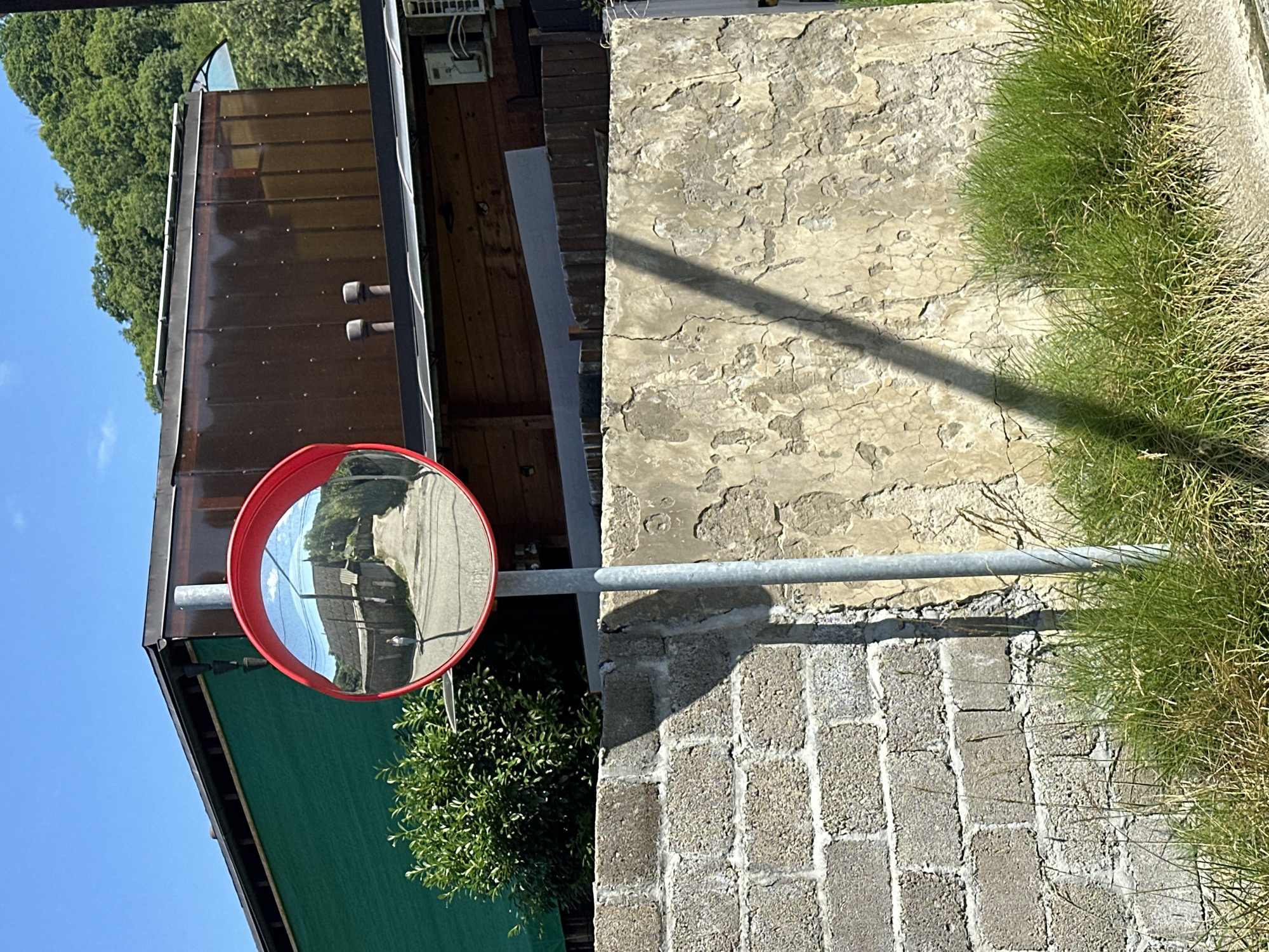 A convex mirror is mounted on a pole in front of a wall. The mirror reflects a narrow street with buildings on either side, a person walking, and green hills in the distance. The background of the image shows a green structure and a brown building. The overall scene is outdoors with a blue sky overhead.