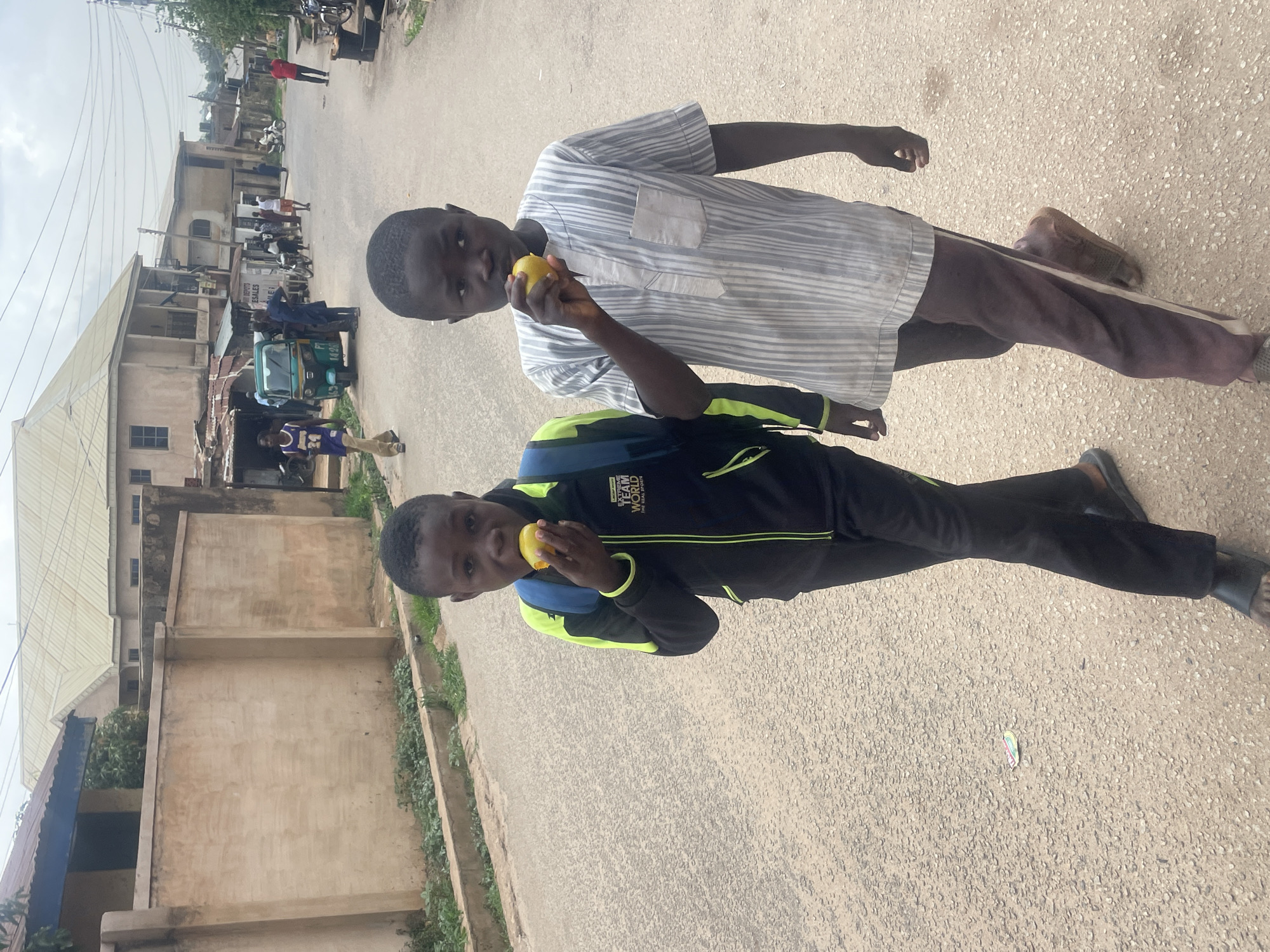 Two boys stand on a paved road in Jos, Nigeria. The boy in front wears a black tracksuit with lime green accents and a blue backpack, while the other boy wears a horizontally striped white and grey shirt and brown pants. Both boys are holding yellow fruits, appearing to be eating them. The background features buildings of varying heights and construction, typical of a city street, including a three-wheeled vehicle and several people in the distance. Overhead wires are visible. The surrounding environment appears to be a residential or commercial area.