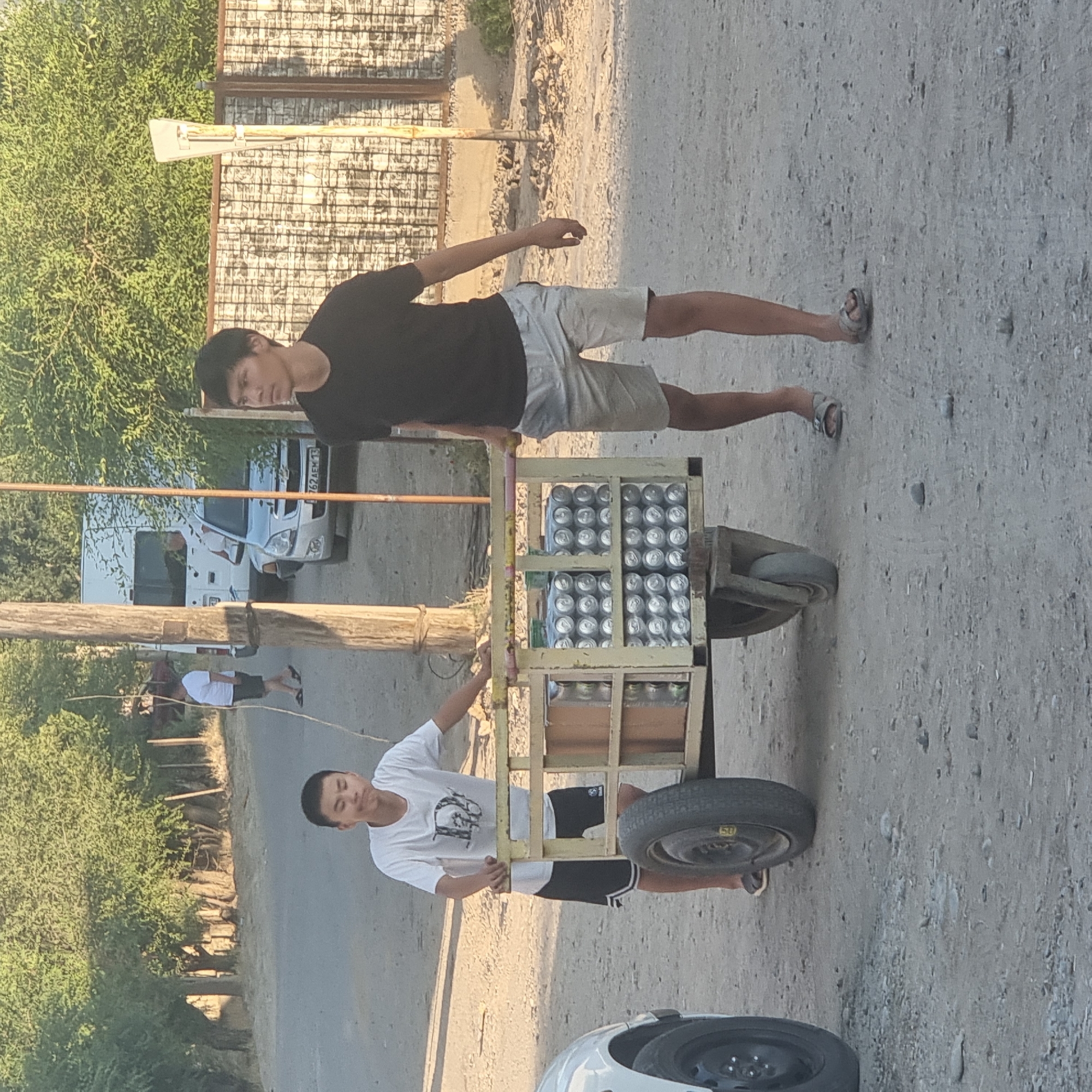 Two young males are pushing a wooden cart loaded with canned beverages down a dirt road in the city of Arys, Kazakhstan. The male on the left, wearing a white t-shirt and black shorts, is positioned behind the cart, with his hands on the side. The male on the right, wearing a black t-shirt and light-colored shorts, is also behind the cart, pushing with both hands. The cart has a yellow frame and is filled with many rows of silver cans. To the left of the cart, a portion of a white car is visible. The background features trees, a white van, a silver car with a license plate that reads "762 AEM 13", and a patterned fence. The ground is a mixture of dirt and gravel. The lighting suggests it is daytime.