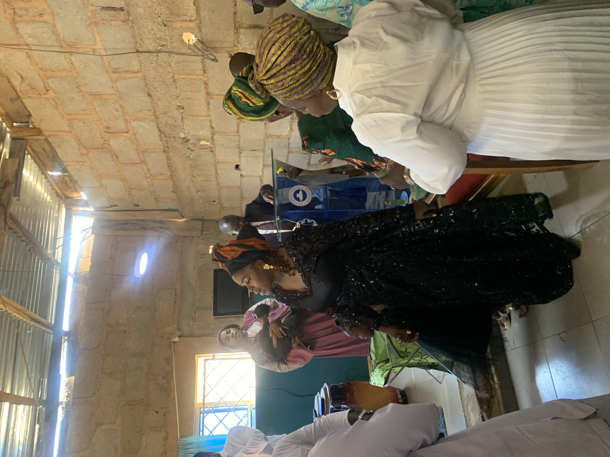 A group of people are gathered inside a building constructed with brick walls and a corrugated metal roof. The location is Gura Top, Nigeria. A woman in a black, sequined dress and orange headwrap stands facing a group, perhaps in a position of leadership or engagement. Another woman, dressed in white, sits with others nearby. There is a podium visible with some writing that is partially blocked in the image. The environment appears to be a place of worship or community gathering.
