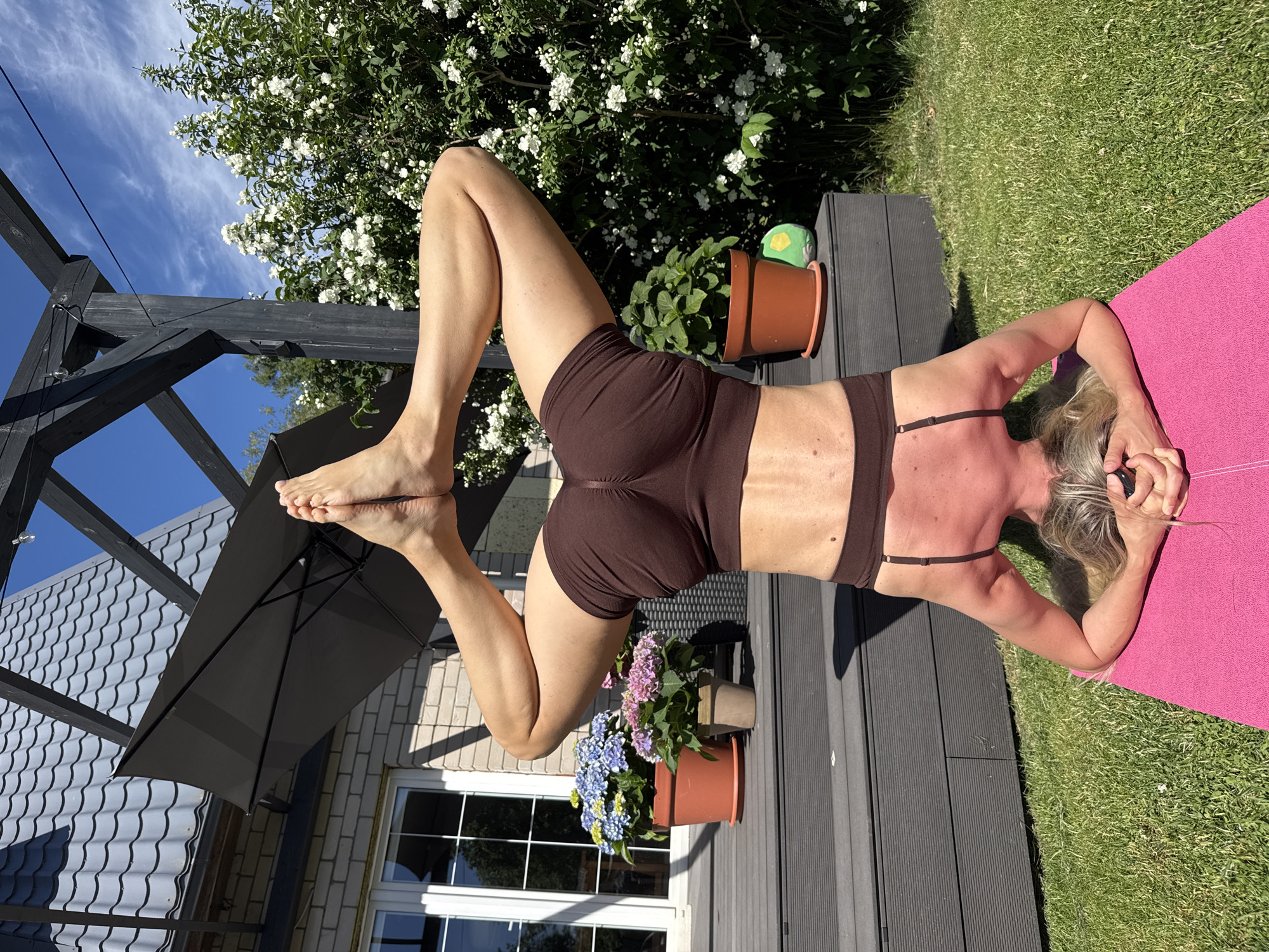 A woman is performing yoga on a pink mat outdoors. She is lying on her back with her legs bent and raised, her feet are crossed. She is wearing brown yoga shorts and a brown sports bra. The background shows a lush green lawn, a wooden deck, a flowering bush with white blossoms, and a clear blue sky with some wispy clouds. A black umbrella is partially visible on the left side of the image, along with part of a house with a grey tiled roof. Two potted plants, one with pink and blue hydrangeas and another with green leaves, are placed on the deck. The scene appears to be set on a sunny day, likely in the late morning or afternoon, given the bright sunlight and clear skies. The woman's posture suggests a focus on flexibility and strength during her yoga practice. The overall impression is one of peaceful outdoor activity and relaxation.