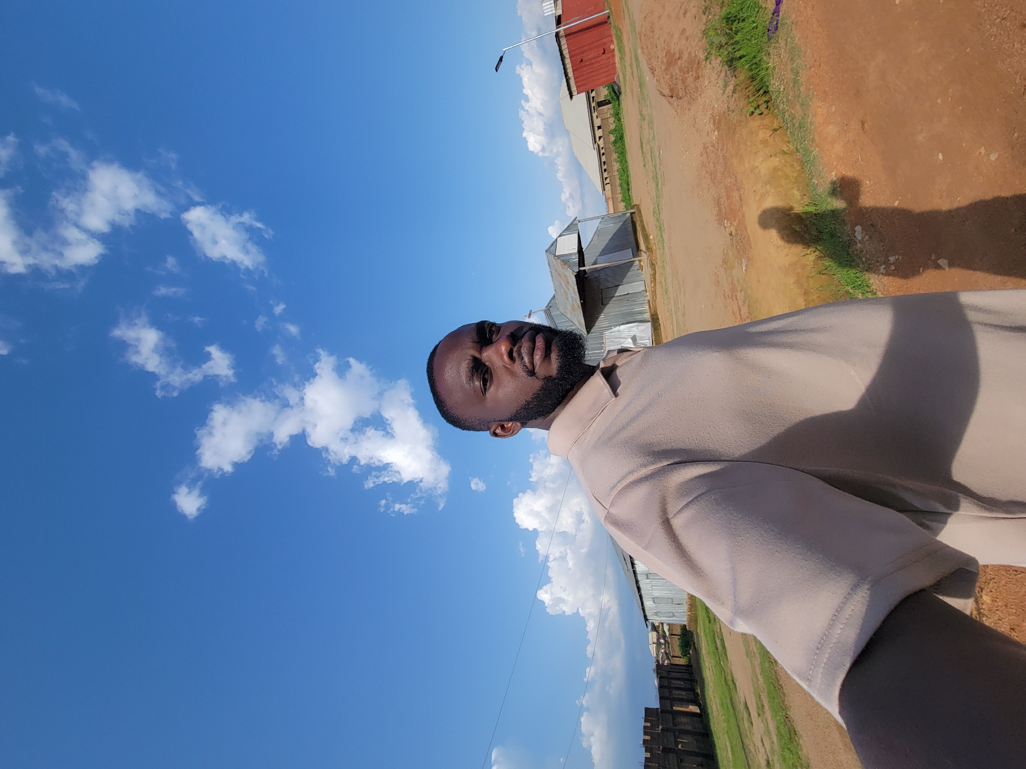 This is a selfie of a Black man with a beard and short hair, wearing a light brown polo shirt. He is standing outdoors with a bright blue sky and scattered white clouds behind him. In the background, there are several structures with corrugated metal roofs, a red building, and a modern street lamp. A fence made of bricks is visible on the left. The ground is reddish-brown dirt with patches of green grass. The lighting suggests it is daytime, possibly late afternoon, given the long shadow of the man cast on the ground. The scene appears to be in a somewhat rural or undeveloped part of the city of Jos, Nigeria, characterized by its informal buildings and unpaved ground.