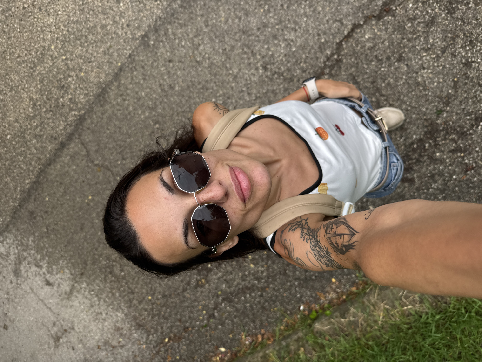 An adult female is captured in a high-angle selfie outdoors, designated as the city of Pleven, Bulgaria. She has dark hair and wears dark, hexagonal-framed sunglasses that reflect the surroundings. Her face shows visible freckles across the nose and cheeks, and her lips are slightly parted in a faint smile. She is wearing a white tank top with black trim, featuring small fruit-themed designs including a pumpkin/orange and a cherry. A light tan strap, likely from a backpack, crosses her right shoulder. Multiple black-ink tattoos are visible on her arms: a large, intricate design on her left upper arm and a stylized sunburst on her right upper arm. A light-colored smartwatch with a red accent is on her left wrist. She is dressed in light blue denim jeans with a tan belt and a portion of a light-colored shoe is visible on her right foot. Her left forearm extends into the lower-left foreground, holding the camera. The background is a textured, light grey asphalt or concrete surface displaying cracks, with patches of green grass and some dry leaves visible along the left edge. The scene is illuminated by natural daylight.