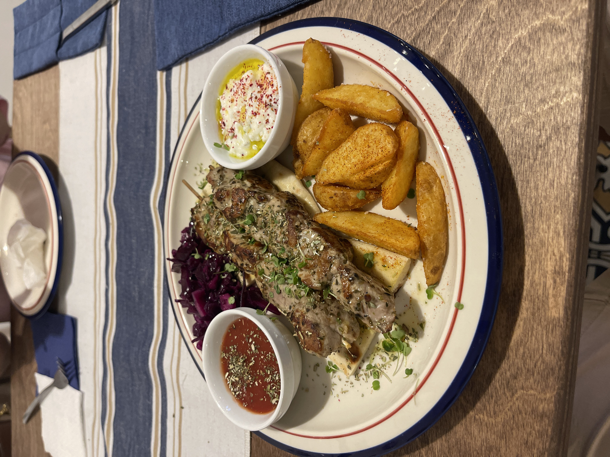 A plate of food sits on a wooden table covered partially with a blue and white striped fabric. The plate, rimmed with blue and red stripes, contains the main course.
The main course appears to be a meat skewer, garnished with herbs and placed atop a bed of two white, rectangular, flatbreads. Alongside the skewer is a serving of cooked red cabbage, a ramekin of red sauce with green herbs, and a ramekin of white cheese topped with red spices and drizzled with oil. Additionally, there are several seasoned potato wedges.
Other table setting elements visible include a white plate, silverware, and a blue napkin.
There is no information from the image to infer the location in Izhevsk, Russia.