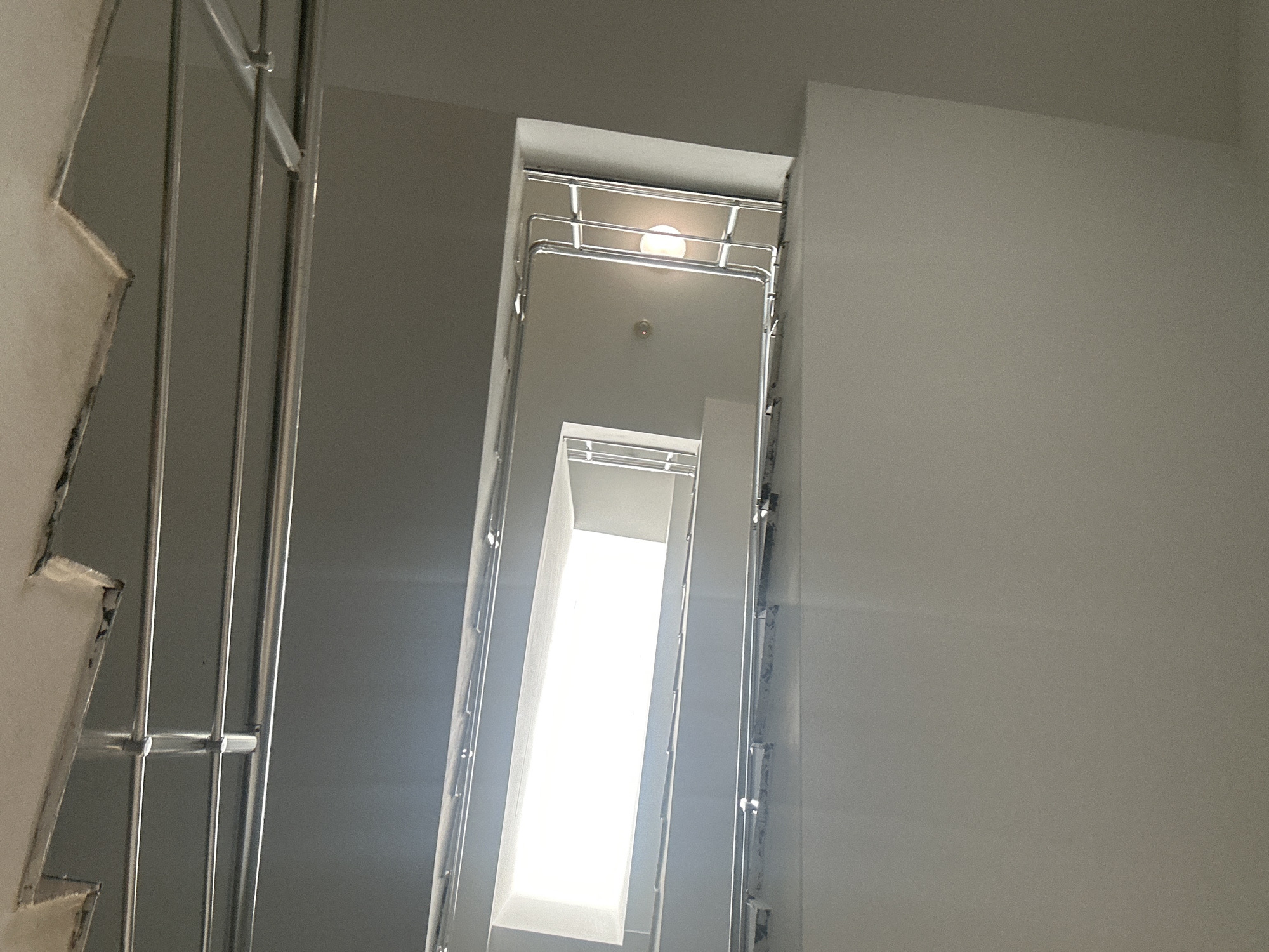The image depicts a view looking up through a stairwell in an indoor environment. Metal railings are visible on the left and along the stairwell walls, ascending upwards. The light source at the top of the stairwell is bright, suggesting a window or a light fixture. The walls are painted in a neutral tone. No people are visible. The location is specified as Unknown, Unknown.