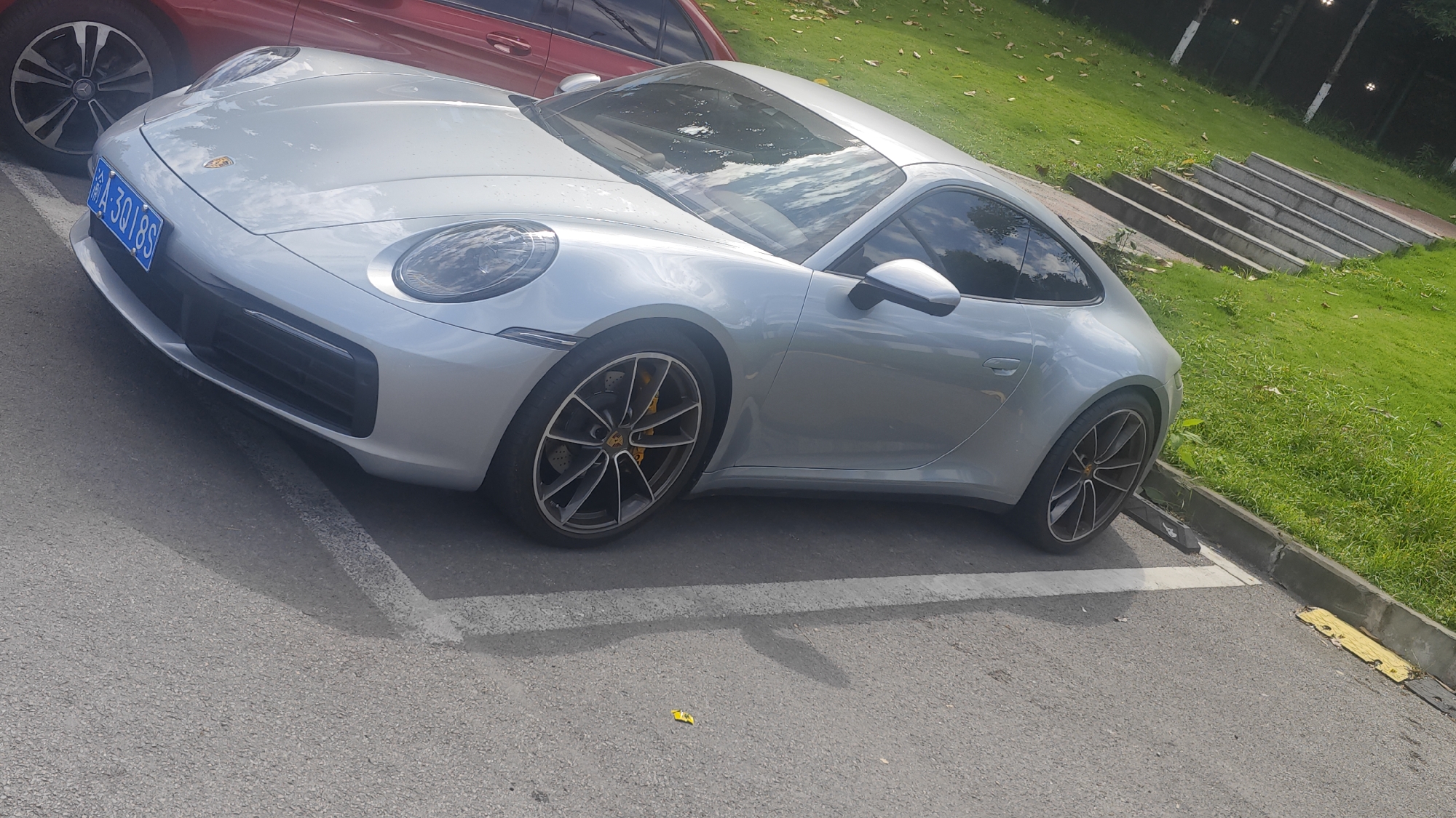The image shows a silver Porsche 911 parked in a parking space on an asphalt surface. The car is parked at an angle within the painted lines. Part of a red vehicle is visible in the upper left corner of the frame. The background includes green grass, some foliage, and what appears to be concrete stairs or a retaining wall. The license plate is visible on the front of the car and appears to be a Chinese license plate (A-3018S). The location is Jiulongpo District, China.
