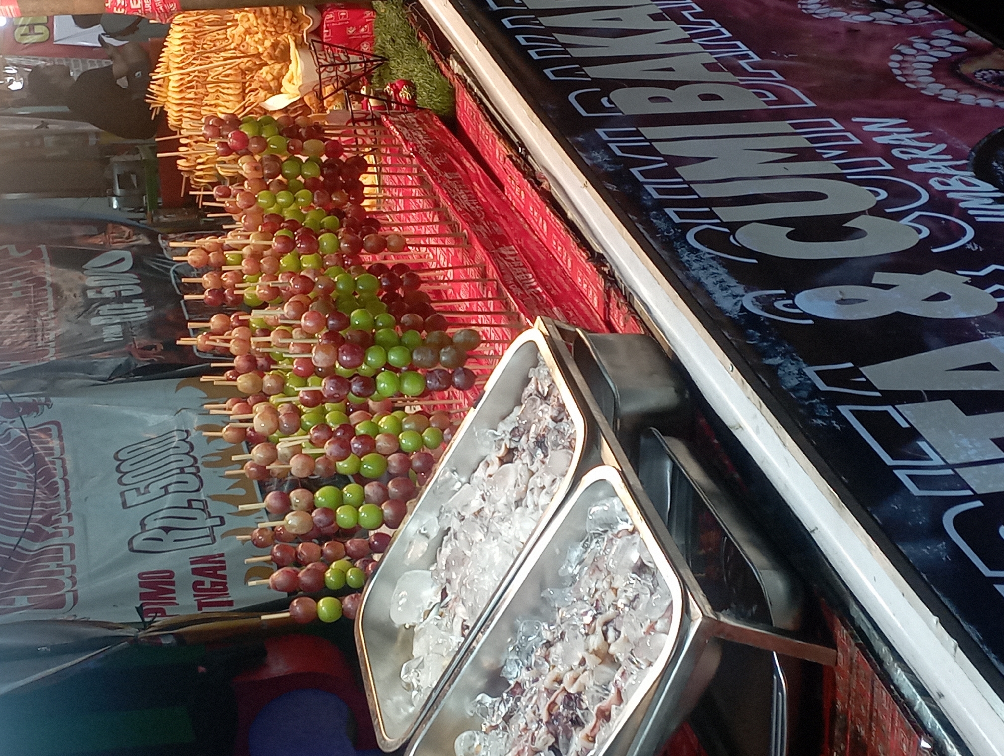 A street food stall in Nganjuk, Indonesia, displays a variety of food items, including skewers of grapes and what appears to be seafood on ice in metal trays. A large banner with Indonesian text and graphics is visible, advertising "CUMI BAKAR" which translates to grilled squid. The stall is illuminated by artificial light, suggesting it is nighttime. The grapes are arranged in colorful rows, with alternating red and green fruit. Behind the grapes, there are more skewers of food, possibly fried items. A person is partially visible in the background, seemingly operating the stall. The overall impression is that of a bustling night market or street food vendor.