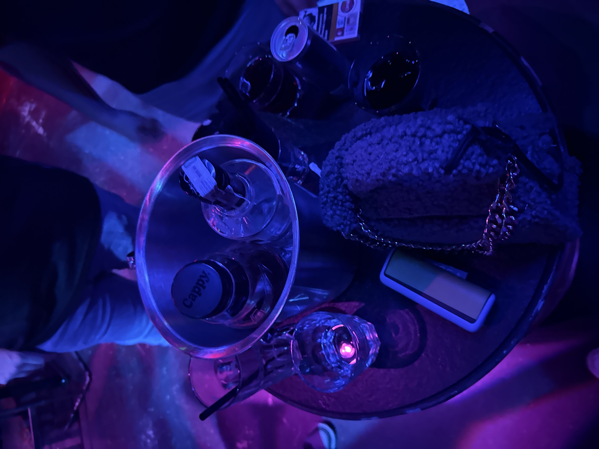 A top-down, eye-level shot under purple and blue lighting captures a circular table in what appears to be a lounge. The table is littered with objects, including open cans of soft drinks, half-filled glasses, a bottle with the label "Cappy," and an unidentifiable bottle in a silver ice bucket. A furry clutch with a gold chain and a rectangular phone are also visible. The edges of people’s clothing and limbs are visible around the table, suggesting a gathering of individuals in a night-time setting. The location cannot be determined as Varna, Bulgaria.