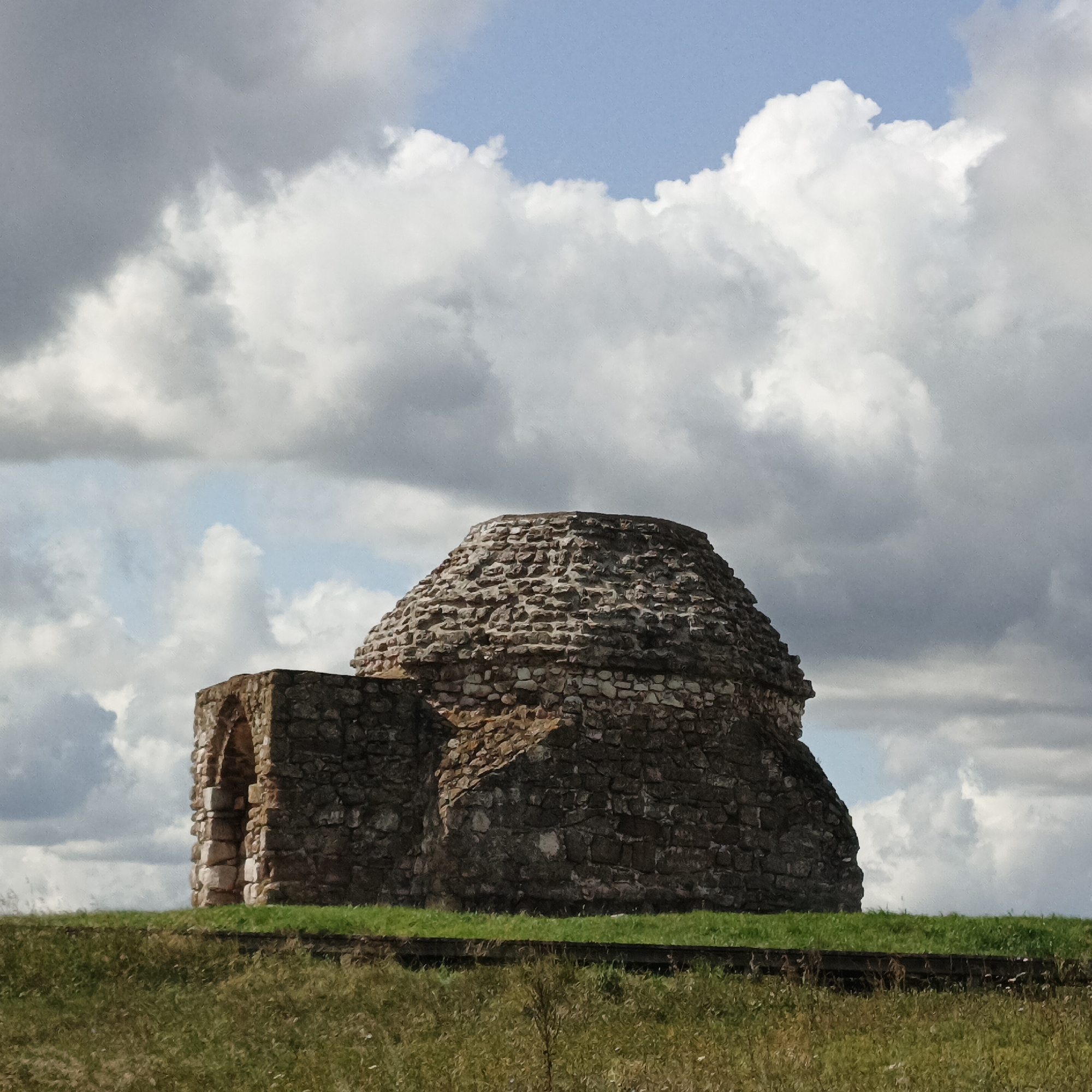 A stone structure with a conical roof and an arched entrance stands on a grassy hill under a cloudy sky. The structure appears to be ancient, constructed from rough, uneven stones. The archway is dark and recessed, hinting at a passage or interior space. The hill is covered in green grass, with some dry vegetation in the foreground. The sky is filled with dramatic, billowy clouds, with patches of blue visible. This scene is located in the vicinity of Verkhnie Termy, Russia, likely at an archaeological site. There are no people or animals visible in the image.