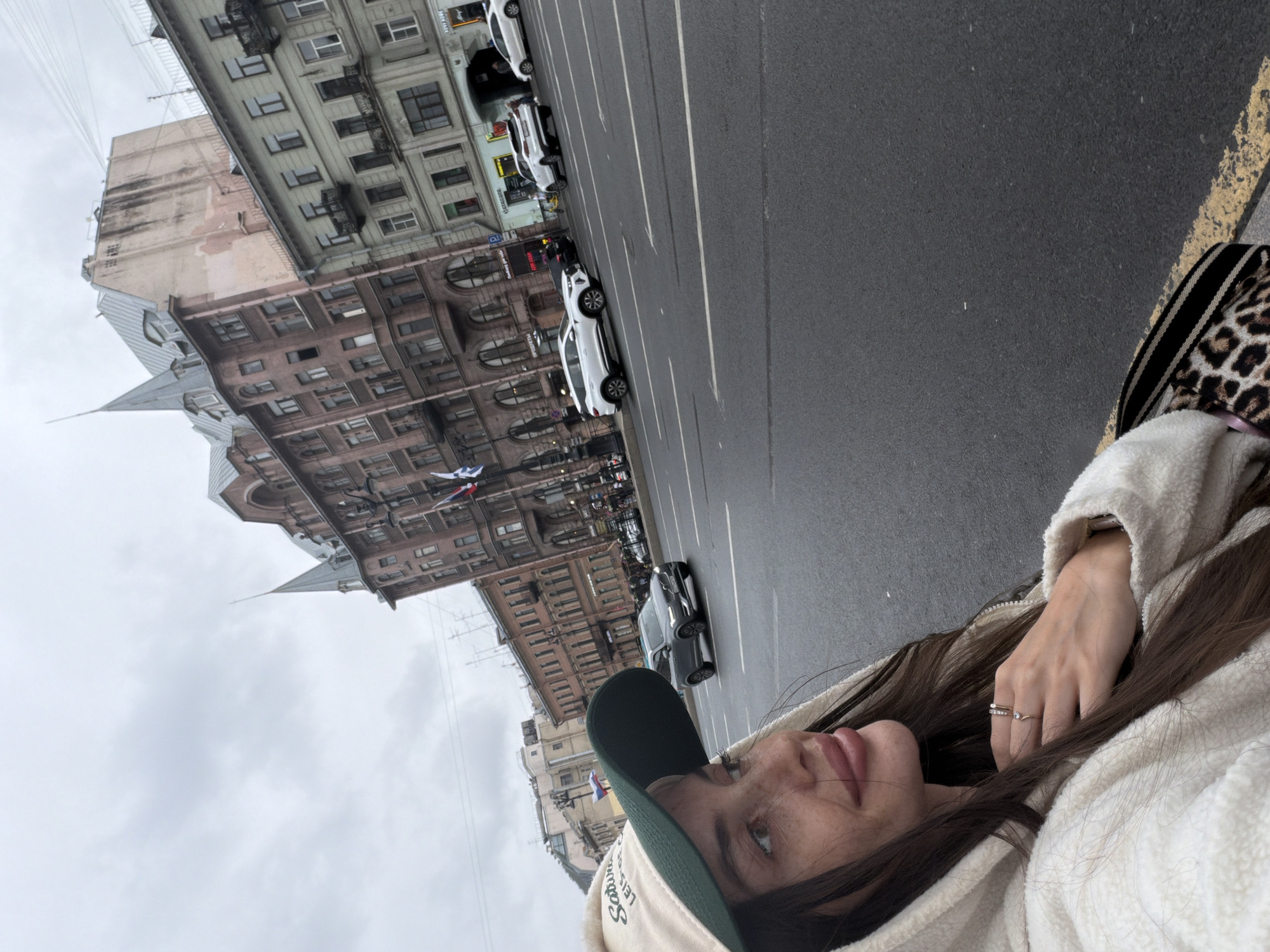 A woman wearing a cream-colored hooded jacket and a tan and green baseball cap is captured in a medium shot, looking to her left. She has long brown hair, visible freckles on her face, and is wearing rings on her left hand. Behind her, a street in Saint Petersburg, Russia, is depicted with several cars on the road, including a grey SUV in the middle lane and white SUVs parked on the right. The street is lined with buildings, some of which have ornate architecture. The sky is overcast.