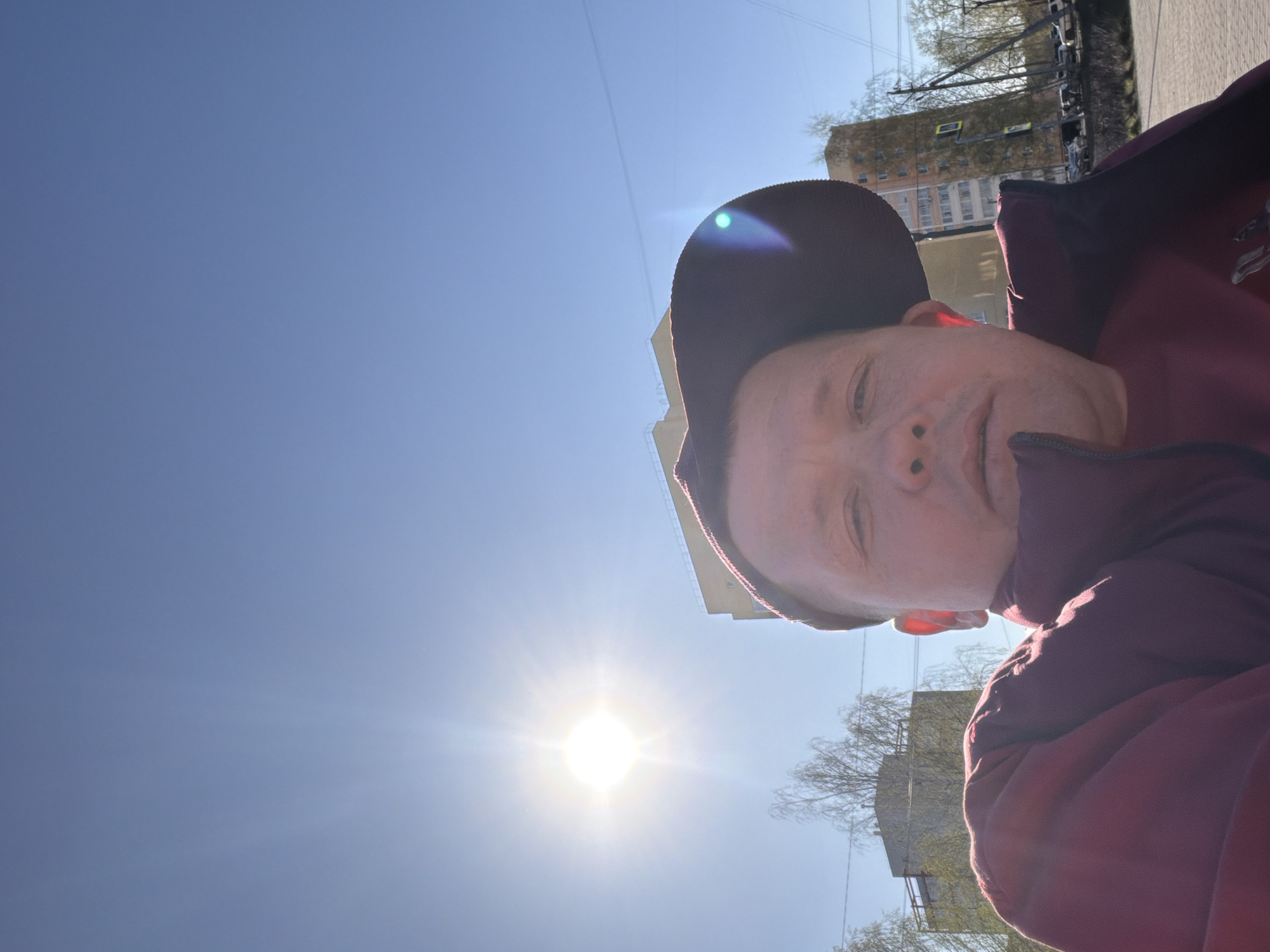 A selfie of a person wearing a dark red hat and jacket taken outdoors on a sunny day. The sun is shining brightly and dominates the lower left portion of the frame. The person’s face is positioned towards the right side of the image. The background shows buildings and trees, suggesting an urban environment in the city of Chaikovsky, Russia.