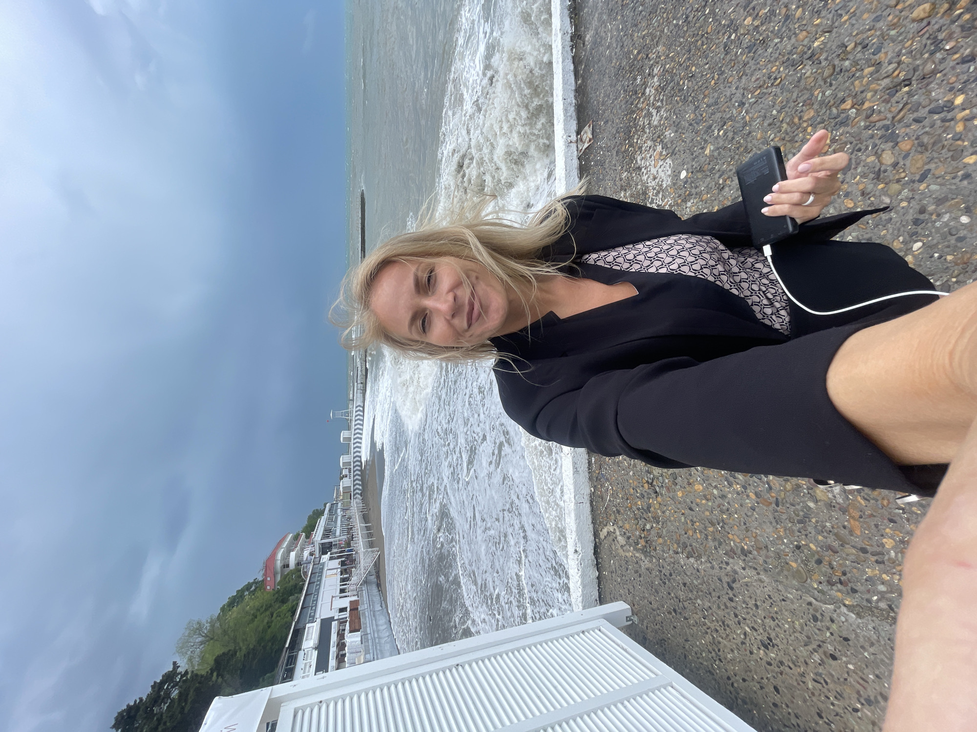 A woman takes a selfie on a stone promenade with the sea crashing against it, likely in Sochi, Russia. She has blonde hair and is wearing a black blazer over a patterned top. She holds a black phone connected to a white charging cord. The sky is overcast. In the background, there are white buildings and trees on a hillside, leading down to the beach. A breakwater extends into the sea.