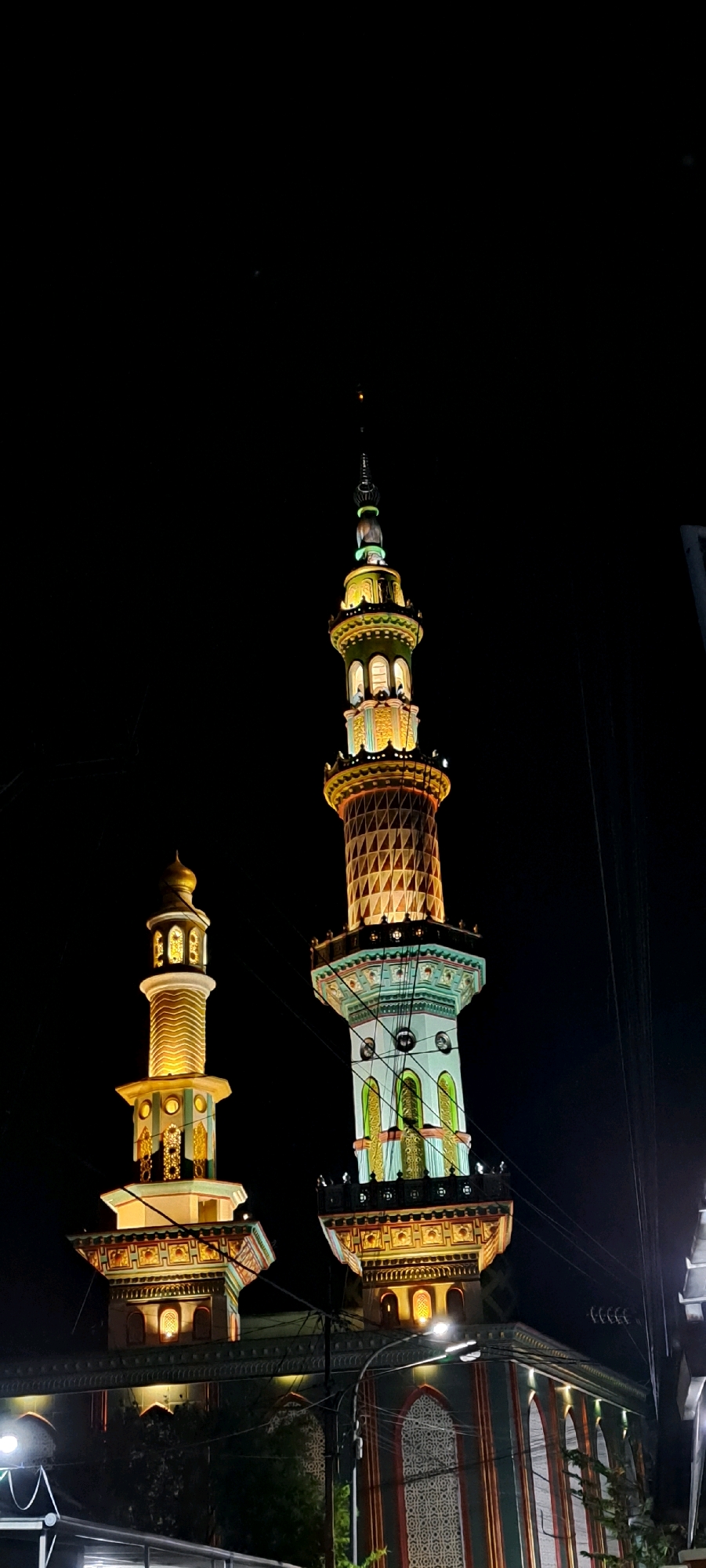This is a vertical, medium-shot, low-angle nighttime photograph of two minarets and a portion of a mosque. The minarets are illuminated by colorful lights. The minaret on the left is shorter and features a golden dome and a pattern of golden wavy lines. The minaret on the right is taller and has a more complex design, with a pattern of triangular shapes in orange and white, a green band with windows, and a decorative finial. The mosque itself, visible at the bottom of the frame, is dark except for illuminated windows and decorative elements. The sky is completely black, suggesting the photo was taken at night. There are several electrical wires visible in the foreground, crossing in front of the minarets. The location is identified as Kebon Waris, Indonesia.