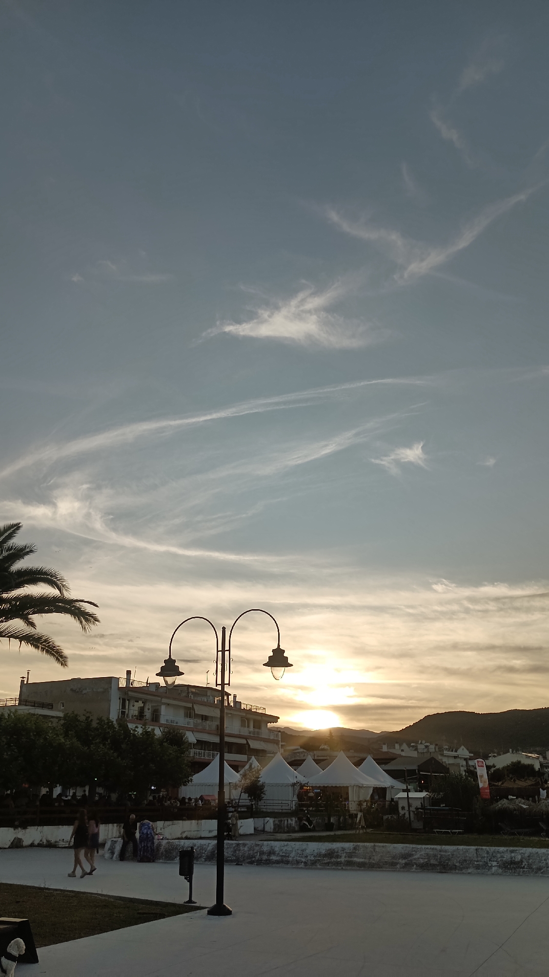 The image depicts a sunset scene in Nea Irakleitsa, Greece, near the waterfront. The sky is a pale blue with wispy white clouds, and the sun is setting behind rolling hills in the distance, casting a warm glow. In the foreground, a black lamppost with two lamps stands prominently, with a paved area in front of it. To the left, a palm tree's fronds are silhouetted against the sky. Behind the lamppost, a row of white event tents are set up, with buildings and more structures visible further back. Several people are walking and standing in the mid-ground, some near the tents and others on a raised walkway lined with trees. The overall atmosphere suggests a relaxed evening by the sea.