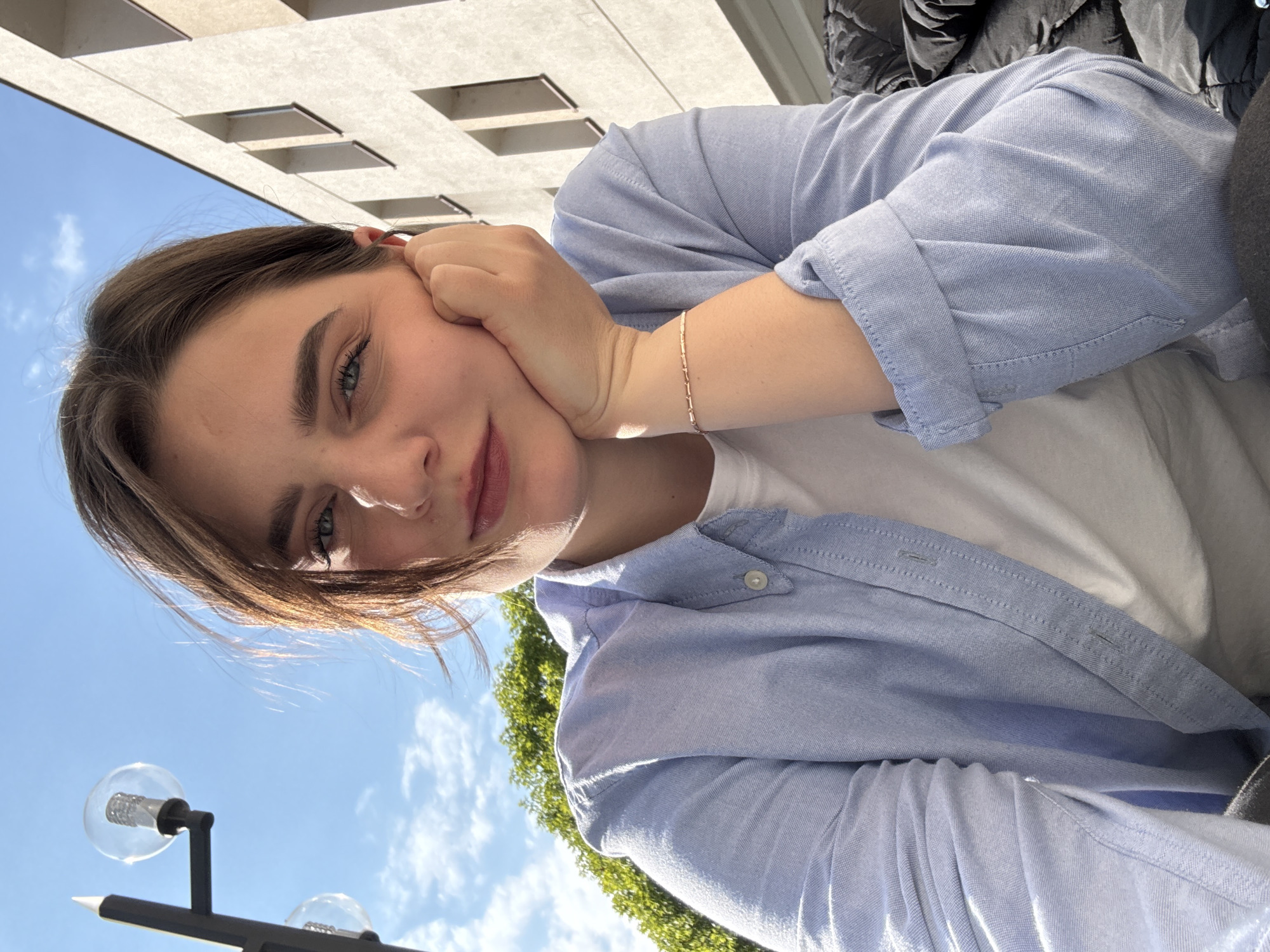 This eye-level, tilted close-up shot features a young individual outdoors. The subject, with medium-length light brown hair and expressive blue-green eyes, gazes directly at the camera with a relaxed, subtle smile. Their right hand rests gently against their cheek, fingers lightly touching their temple. They are dressed in a light blue, long-sleeved button-up shirt with rolled-up sleeves, worn over a white t-shirt. A delicate, rose gold-colored bracelet with small rectangular links adorns their right wrist.
The background is a blend of urban architecture and natural scenery. A light-colored building with dark, rectangular window-like features is visible in the upper portion of the frame, set against a bright blue sky. Lush green tree foliage appears in the lower left, also under the clear blue sky dotted with scattered white clouds. A modern black lamppost with two spherical, clear glass bulbs is partially visible in the bottom left corner. The bright sunlight and clear sky suggest the photo was taken during the day, likely late morning or afternoon. The overall mood is casual and serene, with the individual appearing content. No text is visible in the image. The location context is Trnava, Slovakia.
