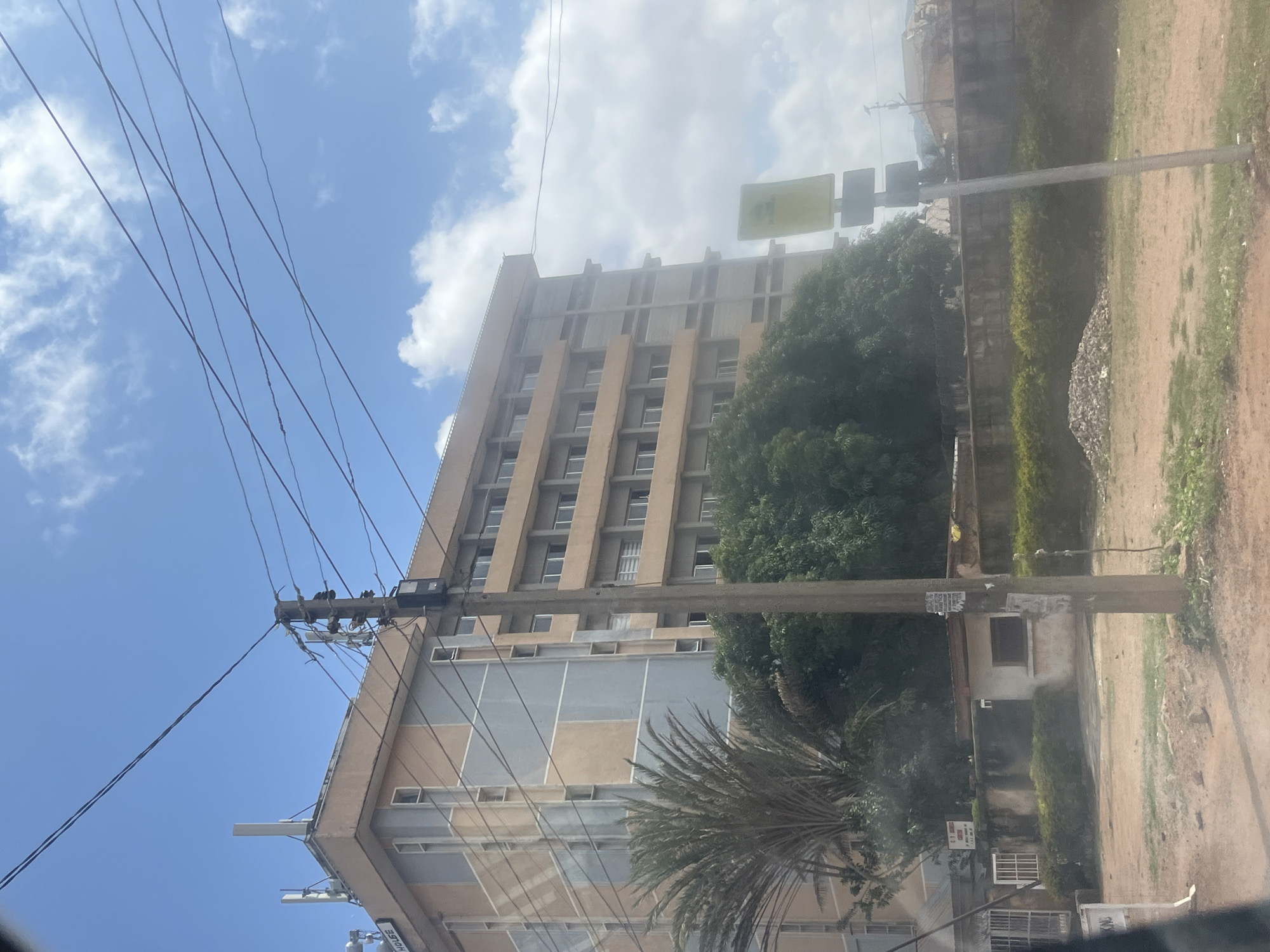 The image depicts a multi-story building located in Jos, Nigeria, viewed from a street-level perspective. The building features a facade with a combination of beige and light blue panels, with multiple rows of windows on each floor. A prominent utility pole with numerous wires stands in the foreground, obscuring parts of the building and the sky. To the left of the building, a palm tree and dense green foliage are visible. In the lower portion of the image, a dirt road or unpaved area is present, with a low concrete wall and some vegetation bordering the building. A yellow traffic or informational sign is mounted on a pole to the right of the building. The sky is partly cloudy with a bright, daytime lighting.