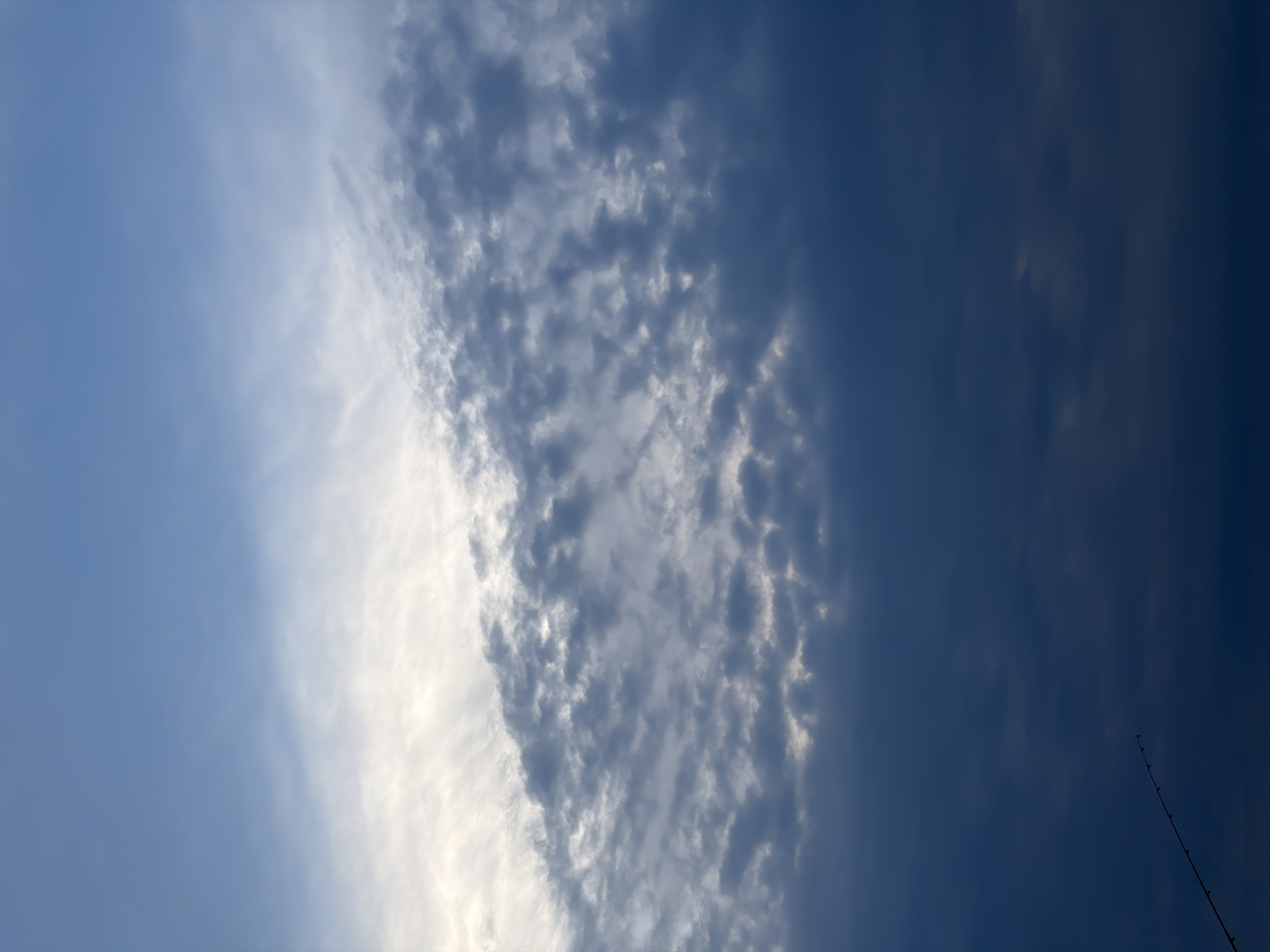 The image primarily features a sky filled with diverse cloud formations and varying shades of blue. On the left, the sky is a soft, light blue with sparse, wispy white clouds. This transitions into a prominent, bright white band of denser clouds running diagonally across the middle of the frame. To the right of this band, the sky darkens considerably to a deep, intense blue, where scattered, darker, and more textured cloud patches are visible. The overall impression is of a partially cloudy daytime sky.
In the bottom right corner, a slender, dark object resembling a fishing rod extends diagonally upwards into the dark blue sky, suggesting the potential for fishing activity, though no person is depicted. The weather appears to be fair, with visible sunlight illuminating parts of the clouds. There are no people or any discernible text in the image. This scene is located in Chaikovsky, Russia.