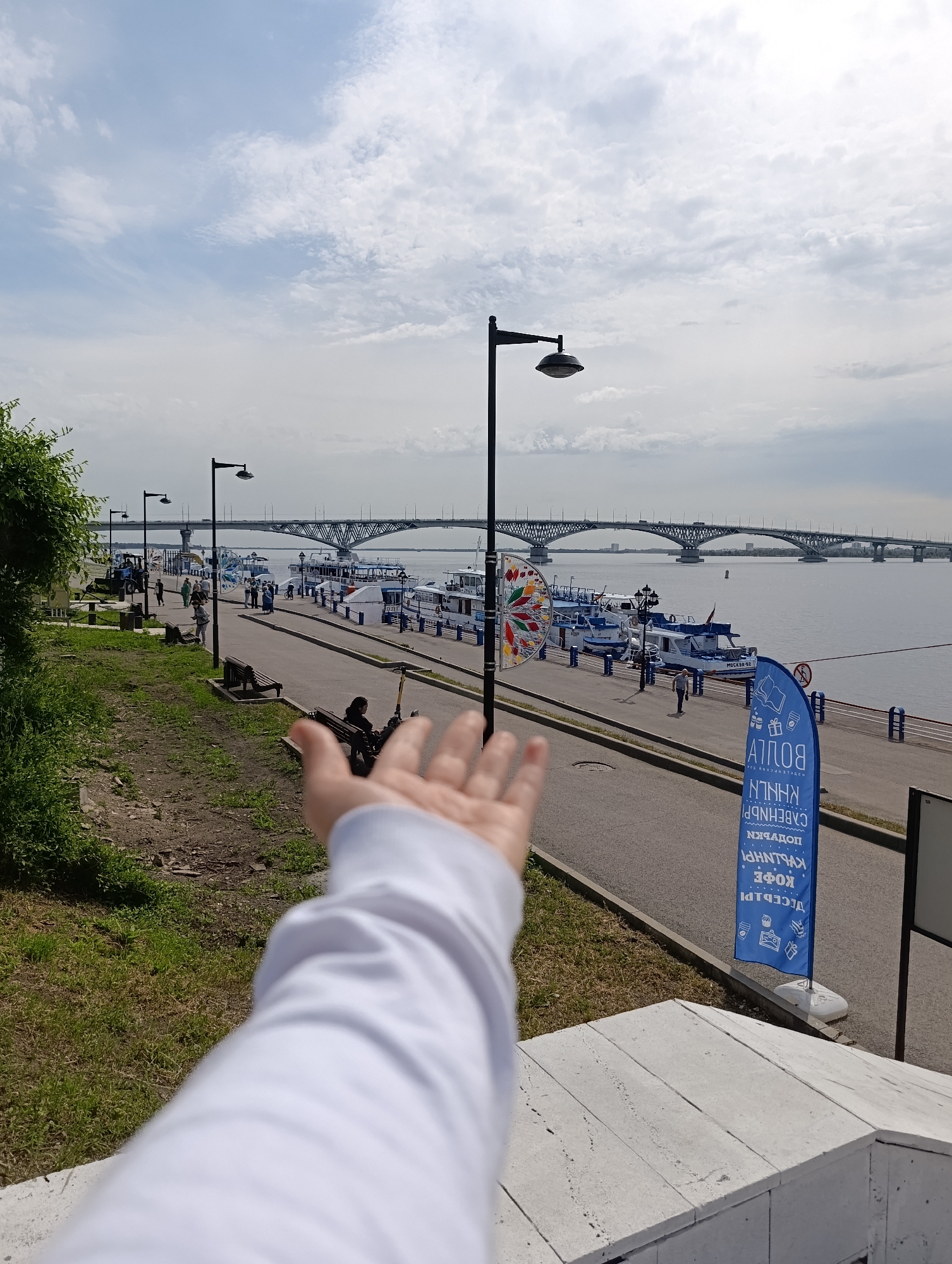The image captures a lively river embankment in Saratov, Russia, under a bright, cloudy sky during daytime. In the immediate foreground, a person's arm, clad in a white long-sleeved shirt, extends towards the scene, as if reaching out or pointing.
The paved walkway along the embankment is populated by several people walking and standing, with benches and streetlights lining the path. On the right, numerous white and blue passenger boats are docked, including one clearly marked "МОСКВА-82" (MOSKVA-82). A colorful, decorative circular ornament on a stand adds a festive element near the boats. Further along, a blue vertical banner, seen inverted, displays Cyrillic text that reads "ВОЛГА" (VOLGA), "САЛЕНДР" (CALENDAR), "ПОДАРКИ" (GIFTS), "КУЛИНАРИЯ" (CULINARY), "КОФЕ" (COFFEE), and "ДЕСЕРТЫ" (DESSERTS), indicating nearby shops or services.
In the background, a wide river stretches under a large, multi-span arch-truss bridge. The left side of the scene features a grassy slope with some green foliage. The overall atmosphere suggests a pleasant, recreational setting, likely a popular spot for leisurely strolls and river activities.
