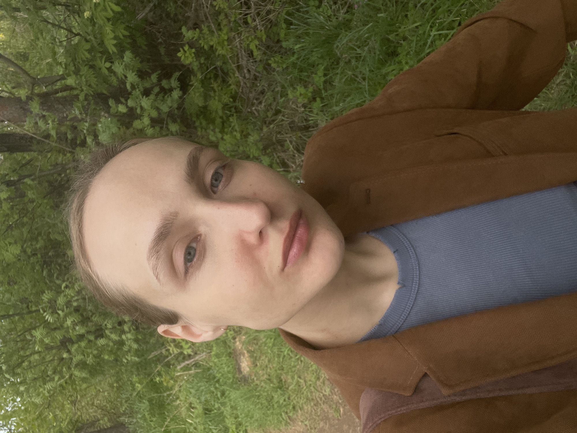 A close-up selfie of a person with fair skin, light brown hair, and blue eyes, wearing a light blue ribbed shirt and a brown suede jacket. The person is situated outdoors, with a backdrop of green foliage, possibly trees and grass, indicating a natural environment. The photo may have been taken in Подшивалово, Russia.
