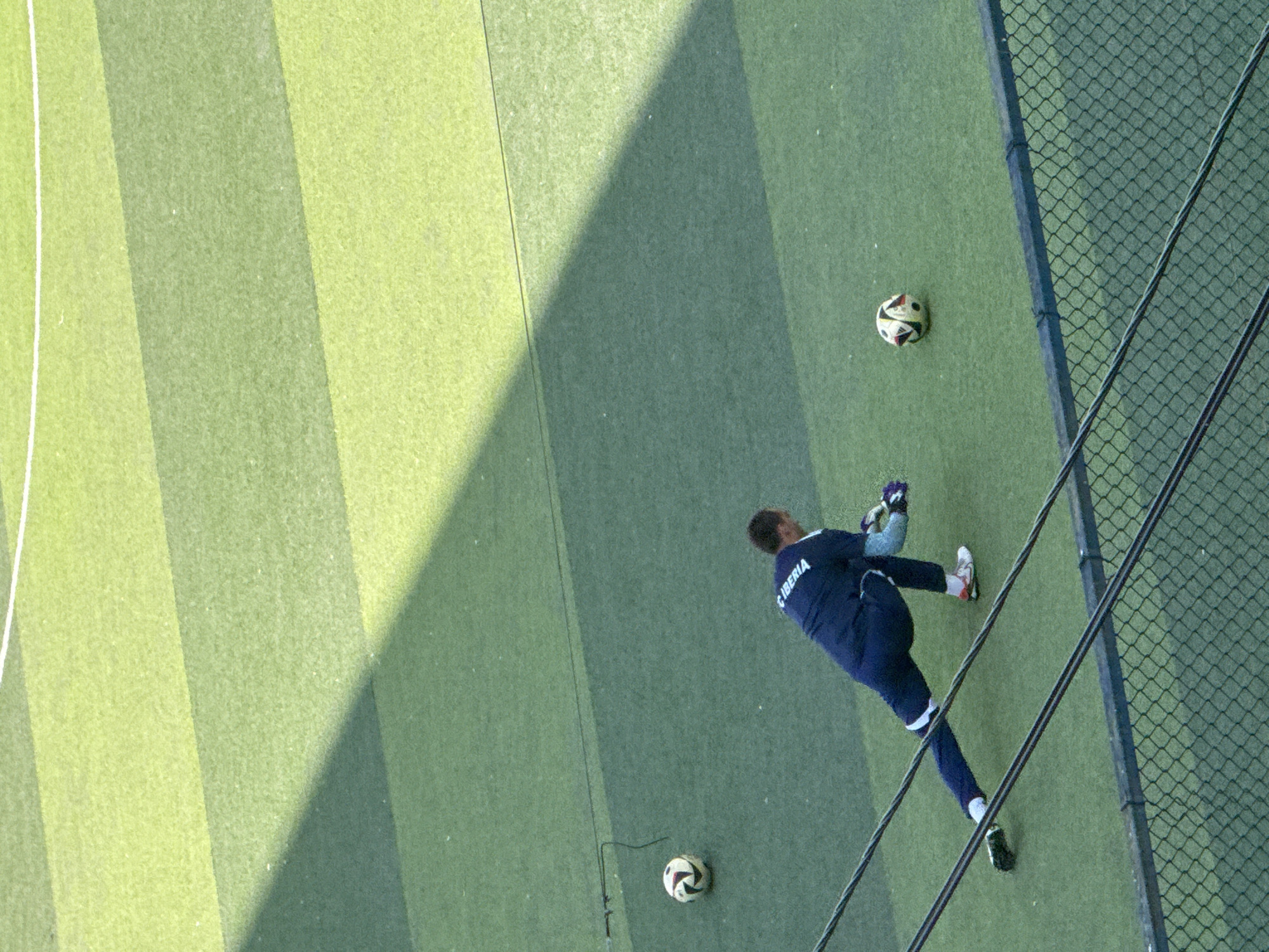 A male soccer goalkeeper in a dark blue uniform with white trim is performing a stretch on a green artificial turf field. He is wearing gloves and is in a low lunge position, with his right leg extended back and his left leg bent forward. A soccer ball is positioned to his left, and another soccer ball is further up the field to his left. The field is marked with alternating shades of green stripes. A black fence is in the foreground, and a shadow from an unseen structure is cast diagonally across the field. This scene appears to be taking place at an outdoor sports facility in Tbilisi, Georgia.