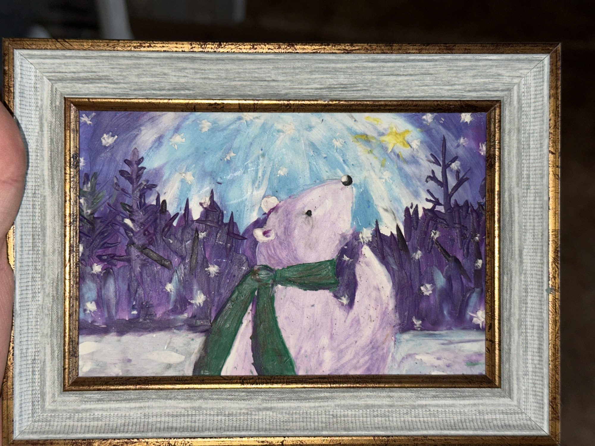 A framed painting depicts a light purple bear wearing a green scarf, standing in a snowy forest under a starry night sky with a bright star. The trees are dark purple, and the painting is framed with a double frame, the inner one being a darker gold and the outer one being light grey. Location: Unknown, Unknown.