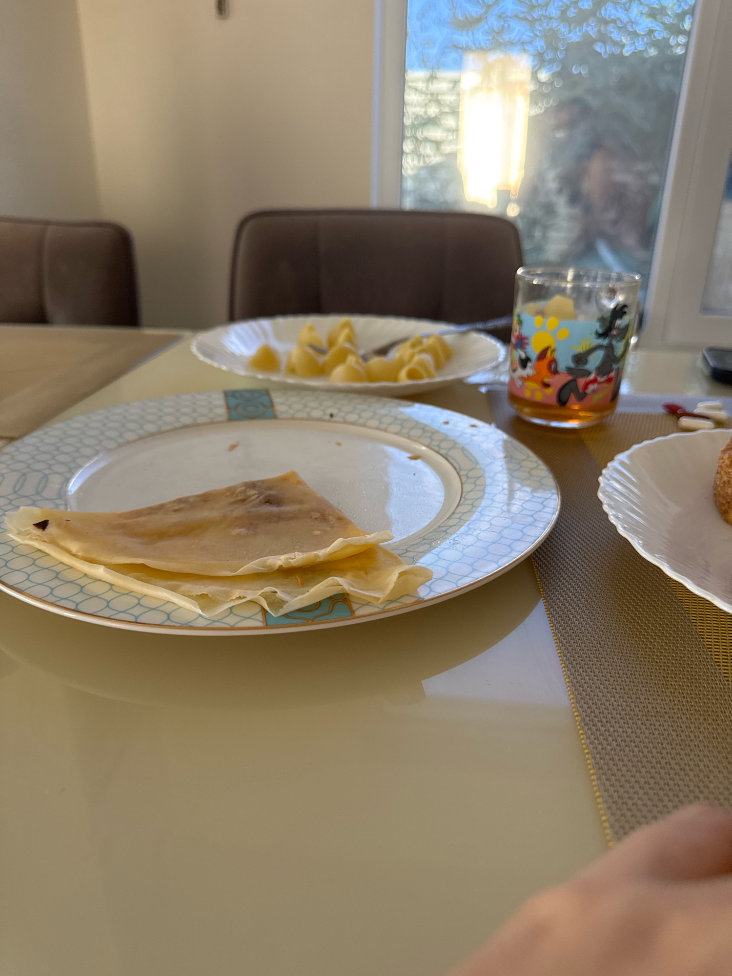A table is set with plates and a glass. In the foreground, a folded crepe is on a white plate with a blue and gold rim. To the right, a small, round pastry is on another white plate. In the background, a glass with cartoon characters is filled with a light-colored beverage. Behind the glass, a plate holds a pile of yellow pasta. Two brown chairs are visible in the blurred background, suggesting a dining setting. The scene appears to be indoors, with light coming from a window. The location specified, Uyutnoye, Ukraine, cannot be determined from the visual information in the image.
