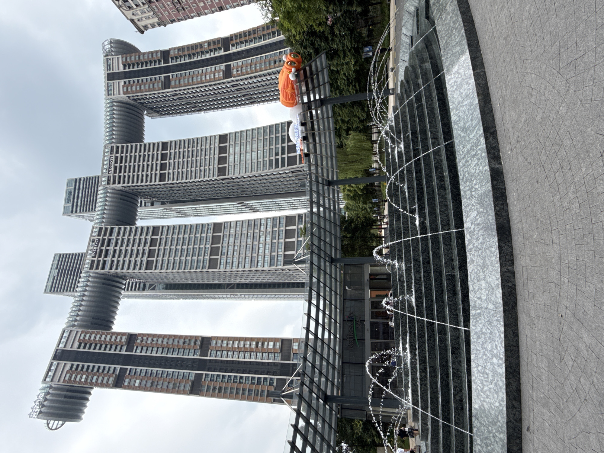 The image shows the Raffles City Chongqing in the Yuzhong District of Chongqing, China. The complex features an iconic structure with multiple towers connected by a horizontal skybridge. In the foreground, a multi-tiered water fountain with several streams of water is active, set against a paved plaza. Behind the fountain, a transparent canopy covers the entrance to a building with the "Raffles City" logo visible. An inflatable orange creature is suspended above the canopy and the building. The sky is overcast. The environment is urban, with trees visible behind the structure and to the right. There are a few individuals visible near the entrance and on the plaza.