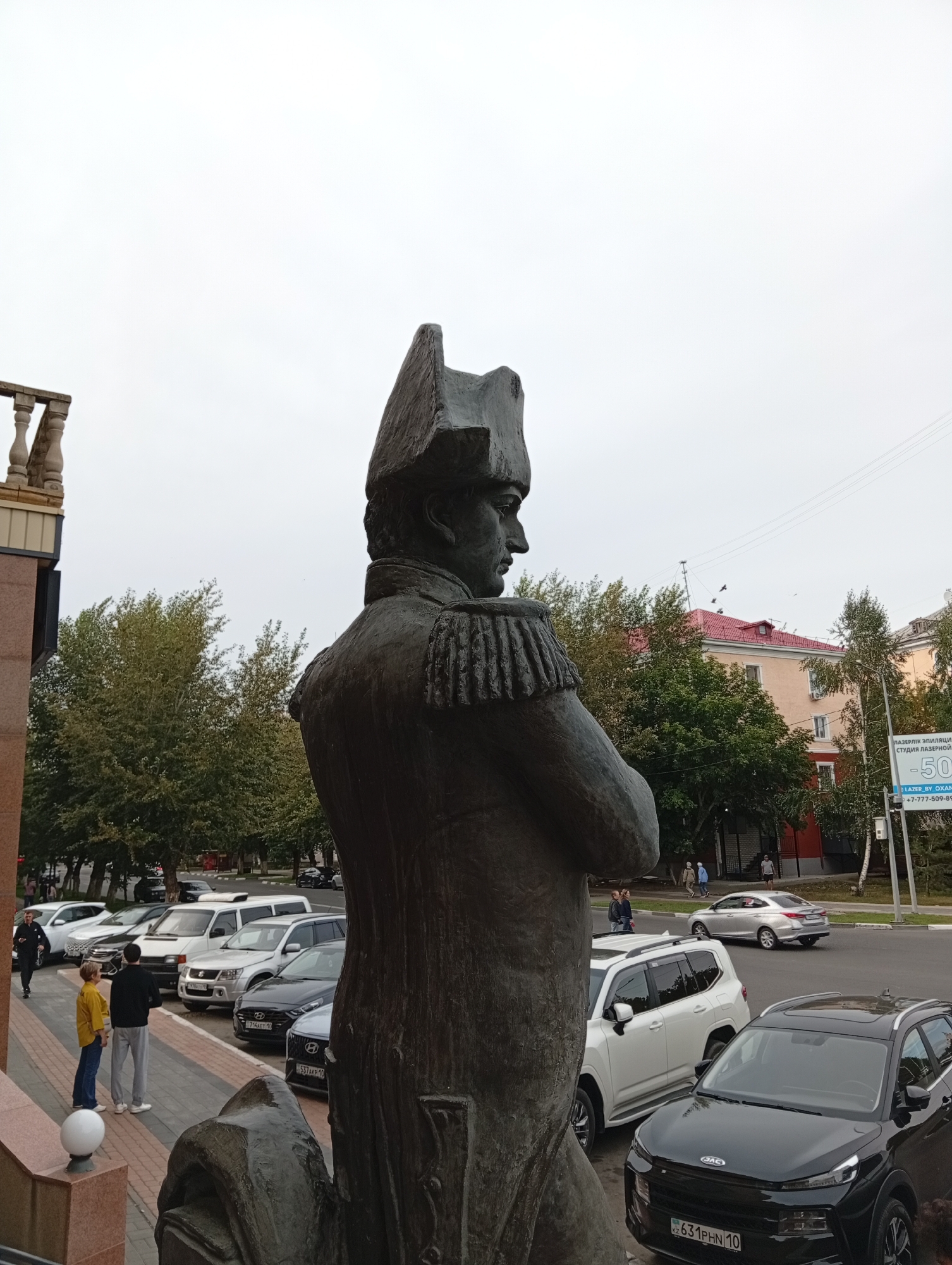 The image shows a statue of a man in a military uniform, facing to the right, with his arms crossed. The statue is made of bronze and is located in the city of Kostanay, Kazakhstan. In the background, there are several cars parked along a street, and some buildings and trees. The sky is overcast.
There are a few people visible in the image. Two people are standing on steps to the left of the statue, looking towards the street. Another person is walking away from the camera to the left. There are also some people walking in the background on the sidewalk.
The statue appears to be in a public space, possibly a square or a park. The surrounding buildings suggest an urban environment. The presence of cars indicates that this is a public street.
The sign on the right side of the image is in Russian and appears to be an advertisement for a laser hair removal studio. It includes the price "-50%" and a phone number.
The overall impression is a typical urban scene with a historical monument.