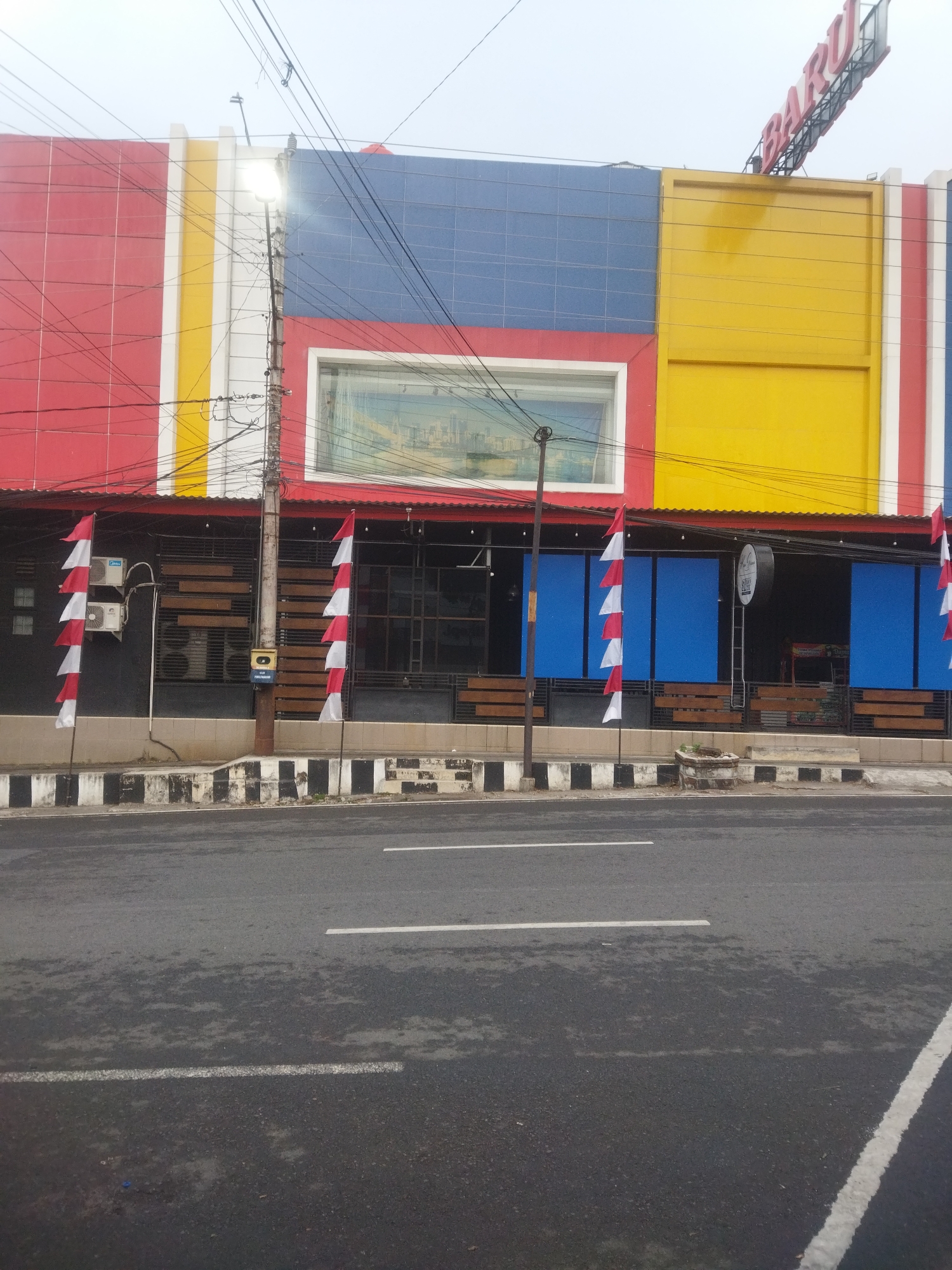The image captures the exterior of a building in Wonogiri, Indonesia, with a vibrant, multi-colored facade. The building features large sections of red, blue, and yellow panels, with white vertical accents. A prominent sign with red lettering that reads "BARU" is mounted at the top right of the building. In front of the building, there is a sidewalk with a distinctive black and white striped pattern. Two poles with Indonesian flags (red and white) are planted along the sidewalk. The building's ground floor has a row of windows and entrances, some covered with blue panels and others with metal grates. Several air conditioning units are visible on the exterior wall. A utility pole with numerous wires and a street lamp stands near the center of the foreground. The street in front of the building is wet, suggesting recent rain. The sky is overcast, and the overall lighting is subdued. There are no people visible in the image.