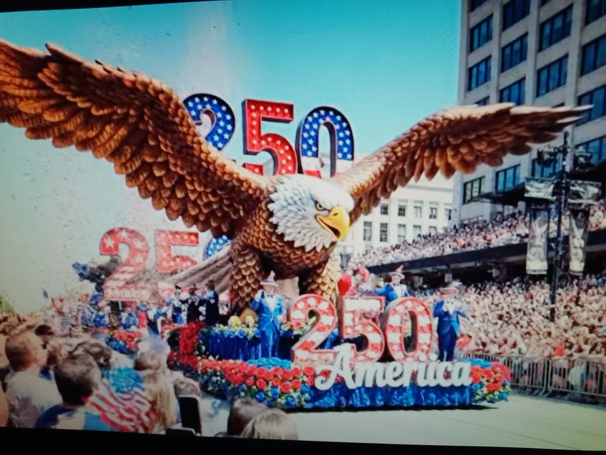 A large, elaborate parade float dominates the scene, featuring a giant eagle with outstretched wings. The eagle's wings are made of numerous brown feathers, and its head is detailed with white and brown plumage and a yellow beak. Behind the eagle, prominent numbers "250" are displayed. The "2" on the left is filled with a blue and white star pattern, while the "50" on the right has red and white striped patterns. Below the numbers, the word "America" is written in white lettering. The float itself is adorned with red, white, and blue floral arrangements and features several figures dressed in blue suits and hats, some appearing to be waving.
In the background, a large crowd of spectators lines the street, with some standing behind barricades. A tall, multi-story building with many windows is visible behind the float and the crowd, suggesting an urban setting. The sky is a clear, bright blue. The overall impression is of a patriotic celebration or parade.
The location is not explicitly provided in the image, and there is no visual information to suggest it is in Chiajna, Romania. The imagery is distinctly American in theme, with the eagle and the word "America."