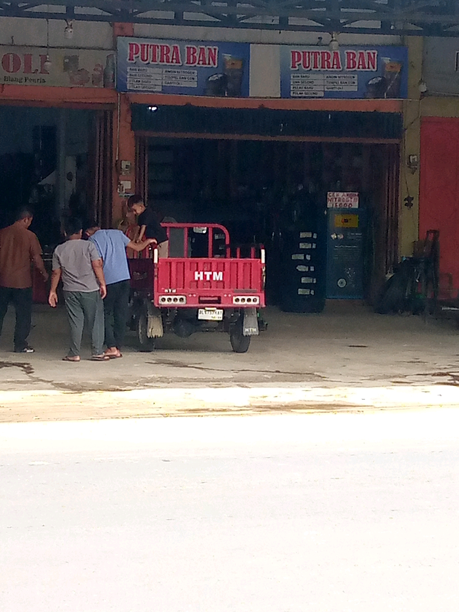 In Keudebayu, Indonesia, a street scene unfolds in front of a commercial establishment. Two men, one in a brown shirt and the other in a gray t-shirt and green pants, stand to the left of a small, red, three-wheeled vehicle. A third man in a blue shirt is standing beside the vehicle, facing away from the camera. A young boy in a black shirt is seated on the back of the red vehicle.
The red vehicle has "HTM" written on its rear. The storefront behind the vehicle displays signs; one reads "PUTRA BAN" in large red letters, and another, partially visible to the left, begins with "OLF". The storefront appears to be an automotive service or tire shop, indicated by the "PUTRA BAN" sign and the presence of tires and automotive equipment in the background. The ground is paved with asphalt, and the overall lighting suggests it is daytime. The environment appears to be urban or semi-urban.