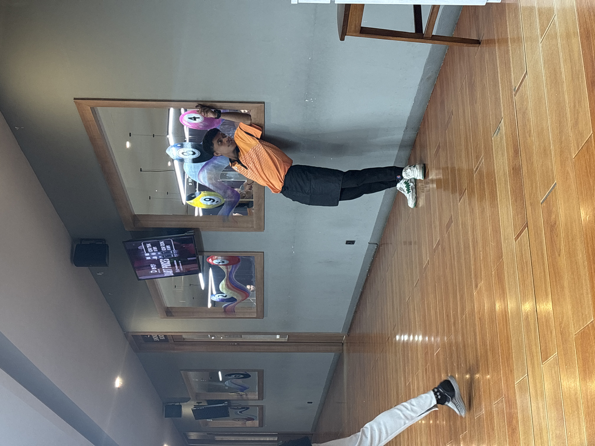 A young man in an orange and black uniform leans against a mirrored wall in what appears to be an indoor entertainment venue, possibly a bar or game room, in Kuta Selatan, Indonesia. The wall reflects brightly colored billiard balls, suggesting a game area. He is wearing black pants and white sneakers. To the left, another person's legs are visible, wearing white pants and similar sneakers, walking away from the camera. The floor is made of polished wooden planks, reflecting the overhead lights. On the wall above the mirrored area, a television screen displays "DART PRICES." The overall environment is dimly lit, with artificial lighting from the ceiling.