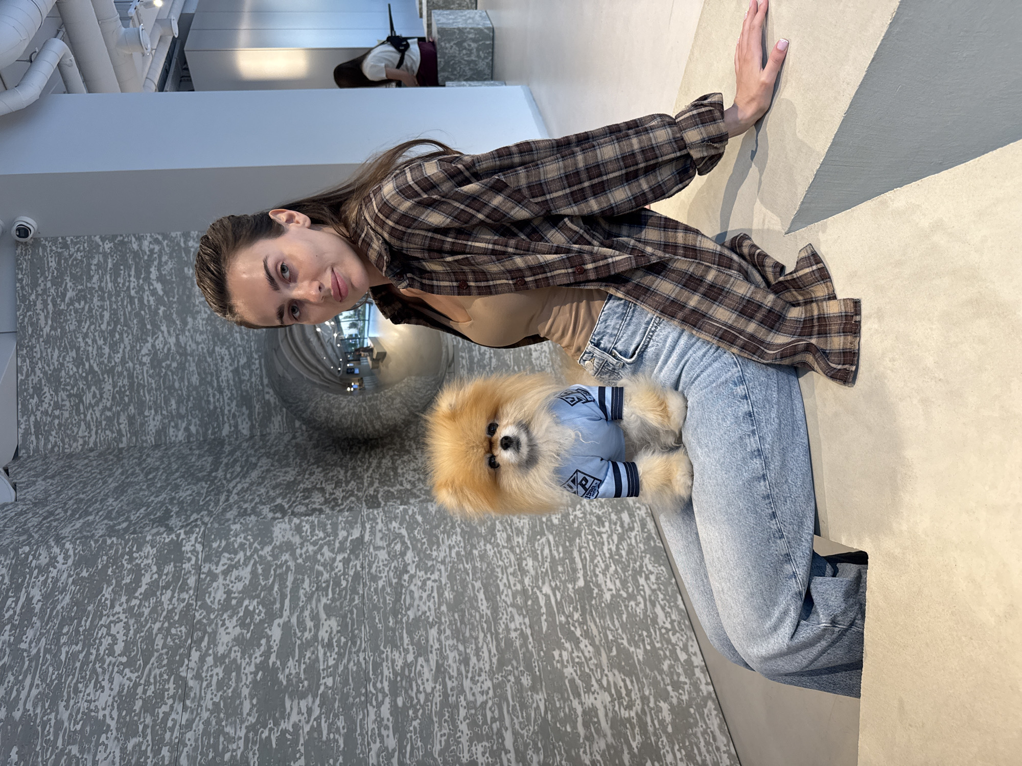 A young woman is depicted sitting indoors, holding a small, fluffy Pomeranian dog. She has brown hair, which appears to be pulled back, and is wearing a brown and tan plaid button-up shirt over a light brown top, paired with light blue jeans. The woman looks directly at the viewer with a calm expression, and her right hand rests on a light-colored, angular surface beside her.
The Pomeranian dog, with light brown fur, black eyes, and a dark nose, is held in the woman's lap. It wears a light blue pet garment featuring dark blue striped sleeves and a crest-like design on the chest. Visible text on the crest includes "DS" and what appears to be "BER" below it. The dog is also looking towards the viewer.
The setting is a brightly lit, modern indoor space. The floor has a distinctive textured pattern of grey and white. In the background, white walls, white ceiling pipes, and a security camera are visible. A large, reflective spherical object can be seen behind the woman, and a blurred figure of another person is present in the distant background. The scene conveys a relaxed and casual moment between the woman and her pet.