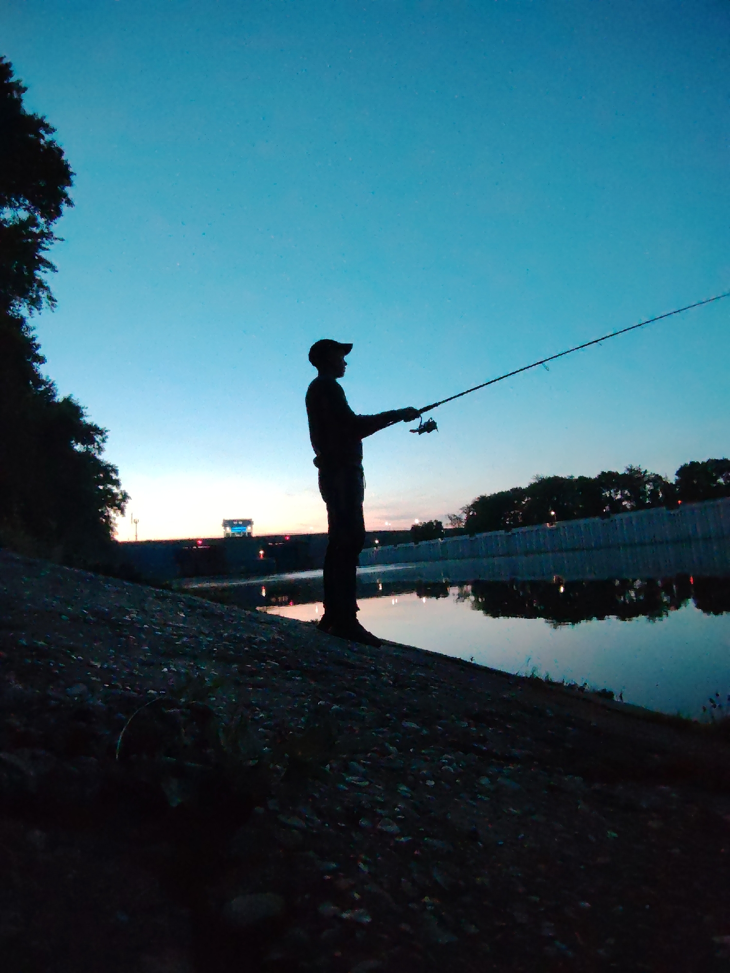 The image shows a silhouette of a person fishing. The individual is wearing a hat and holding a fishing rod. The person is standing on a rocky bank next to a body of water, reflecting the twilight sky. Trees line the bank. In the background, there is a structure with lights. The scene is at dusk or dawn, with a gradient of blue and orange in the sky. The location is likely in the city of Chaikovsky, Russia.