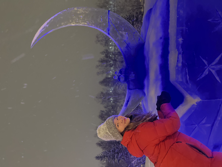 A nighttime scene in Krasnoyarsk, Russia, depicting an outdoor ice sculpture during snowfall. The specific location is likely Theater Square. In the foreground, a female individual, dressed in a red puffer jacket, a light gray knitted beanie, and black gloves, occupies the left side of the frame. She is looking towards the viewer with a slight smile, leaning forward, with her hands clasped at chest height. Her hair is visible beneath her beanie. To her right, a large ice sculpture forms the central subject. It consists of an upright, translucent crescent moon, internally illuminated with blue light, giving it a vibrant blue hue. Two smaller, seated humanoid figures, also carved from ice, are positioned within the lower curve of the crescent moon. The moon sculpture rests on a rectangular ice base, which is also illuminated in blue and features carved star-like patterns on its visible surface. A light layer of snow covers the top surfaces of the ice sculptures. Snowflakes are visibly falling across the entire scene. In the background, a line of snow-covered trees, including conifers and deciduous trees, stretches horizontally. The ground is covered in a uniform layer of snow.